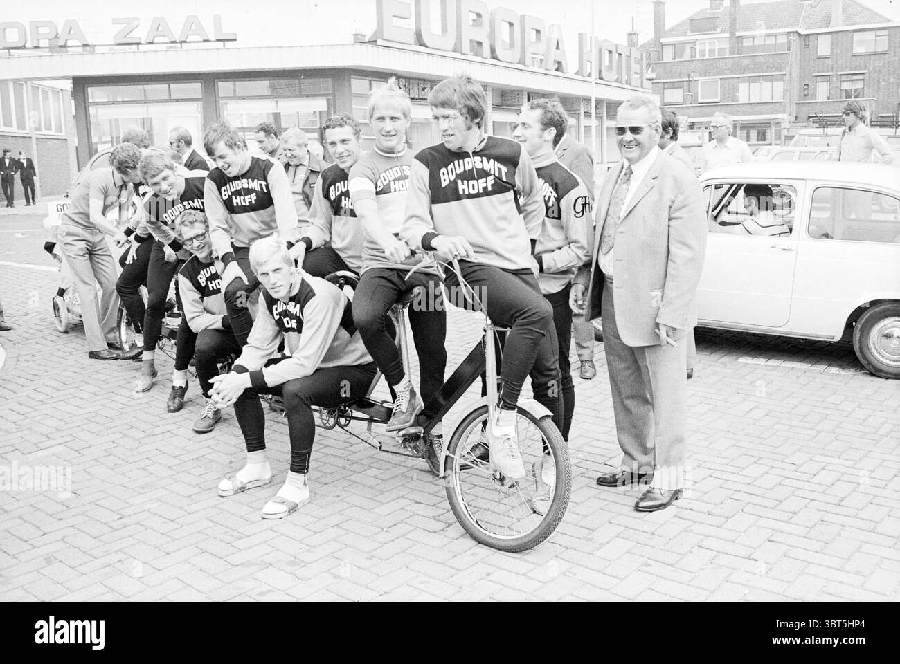 Das Radsportteam Goudsmit Hof Portrait Eddy Merckx porträtiert das belgische Radsportteam Cycling, Whizgle News, Dutch Desk, Niederlande, 1950 - 2000 am 27-06-1972. Diese Themen sind in der Abbildung dargestellt. In einer lebhaften Outdoor-Umgebung wird eine Gruppe von Athleten hervorgehoben, was eine dynamische Szene schafft. Die Einzelpersonen tragen passende Sportkleidung, darunter spezielle Sweatshirts mit einem Namen auf der Vorderseite. Ihre Kleidung zeigt eine Mischung aus Farben, hauptsächlich in Schwarz und einem ergänzenden Farbton, die möglicherweise ihre Teamidentität zum Ausdruck bringen. Einige Mitglieder der Gruppe sitzen auf einem großen, großen Tand Stockfoto