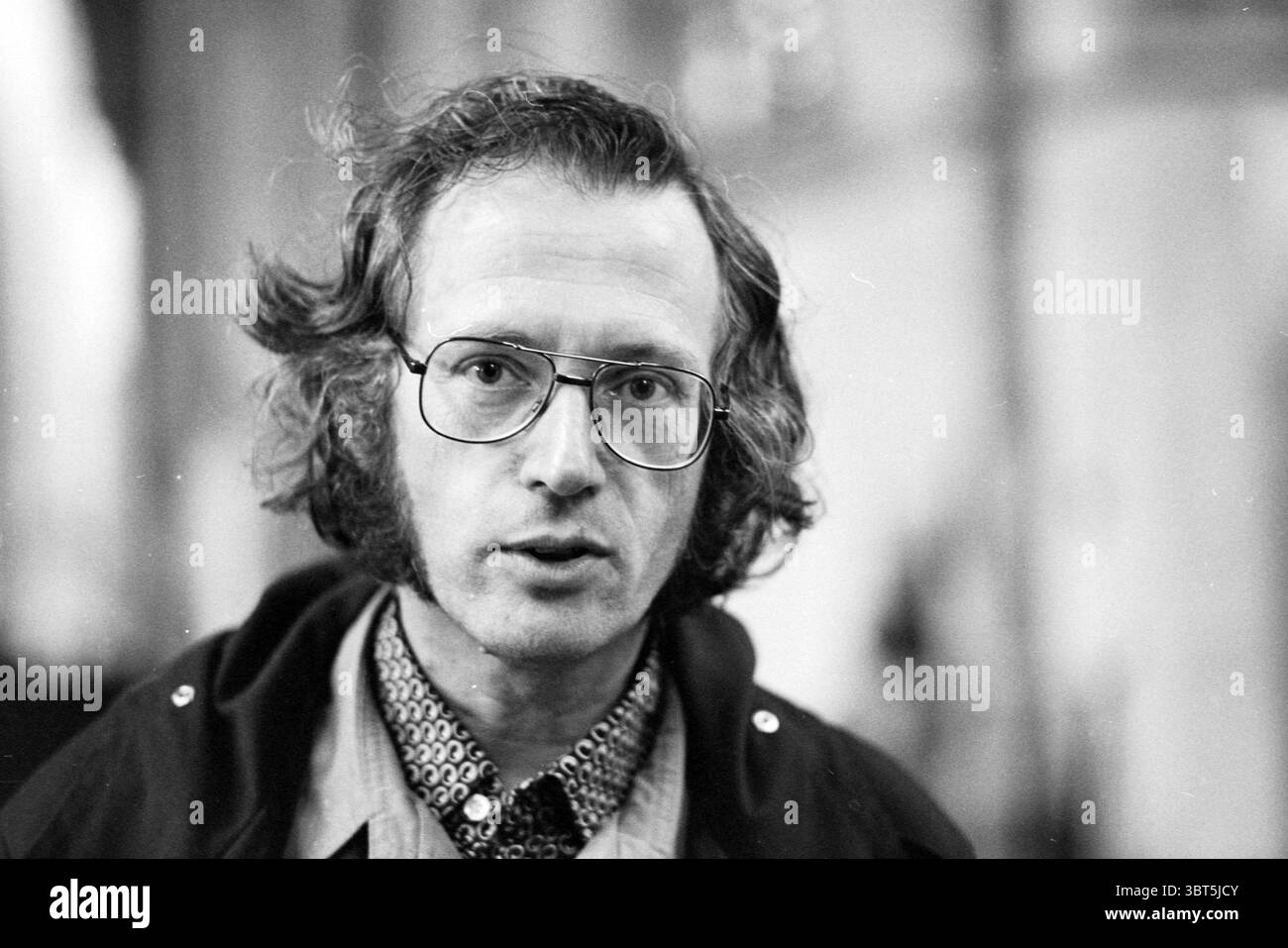 Organist Piet Kee Orgeln und Organisten, Whizgle News, Dutch Desk, Niederlande, 1950 - 2000 am 20-07-1978. Dies sind die Elemente im Bild. Die Szene zeichnet sich durch eine Nahaufnahme eines Individuums mit charakteristischen Merkmalen und einem ausdrucksstarken Verhalten aus. Die Person hat helles, welliges Haar, das das Gesicht umrahmt und zu einem etwas ungezähmten Look beiträgt. Ihre Brille ist auffällig und rund und weist auf einen Vintage-Stil hin, der ihr Gesamtbild ergänzt. Das Motiv trägt ein gemustertes Hemd, das mit einem Design verziert ist, das visuelle Aufmerksamkeit verleiht und unter einem dunklen Mantel getragen wird. Die Kombination aus Stockfoto