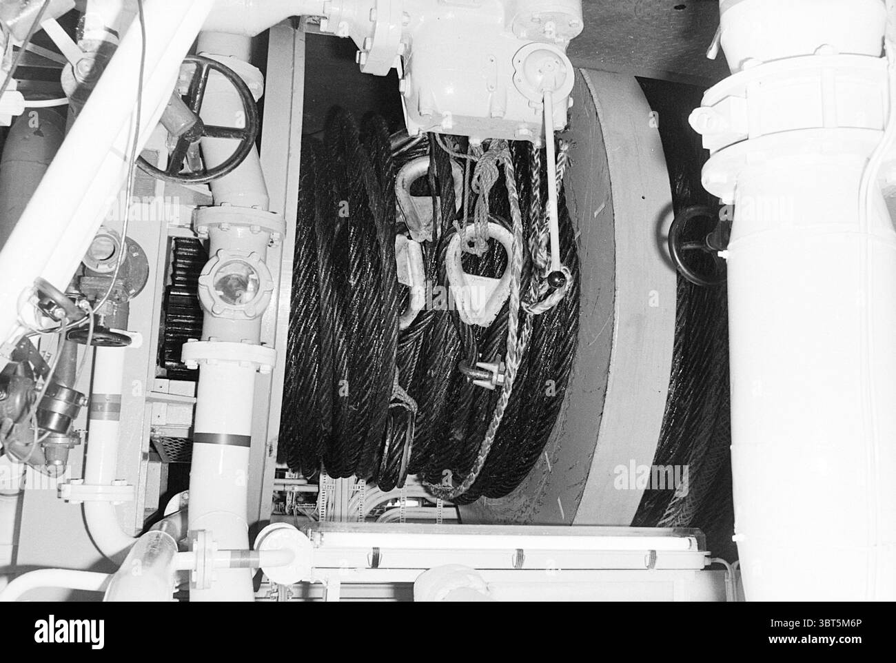 Winde auf Wijsmullers Schlepper., Whizgle News, Dutch Desk, Niederlande, 1950 - 2000. Das Bild enthält diese Themen. Die Szene wird von einer industriellen Umgebung dominiert, die eine große, runde Spule aus dicken, dunklen Kabeln aufweist, die eng gewickelt sind. Die Kabel sind strukturiert und glänzend und werden von einer starken Deckenleuchte beleuchtet, die starke Kontraste von Licht und Schatten erzeugt. Um den Schieber herum befinden sich verschiedene mechanische Komponenten, darunter große Metallräder und -Hebel, die das Gefühl der Funktionalität erhöhen. Die Oberflächen sind metallisch und poliert, reflektieren die Beleuchtung und betonen die Utilitaria Stockfoto