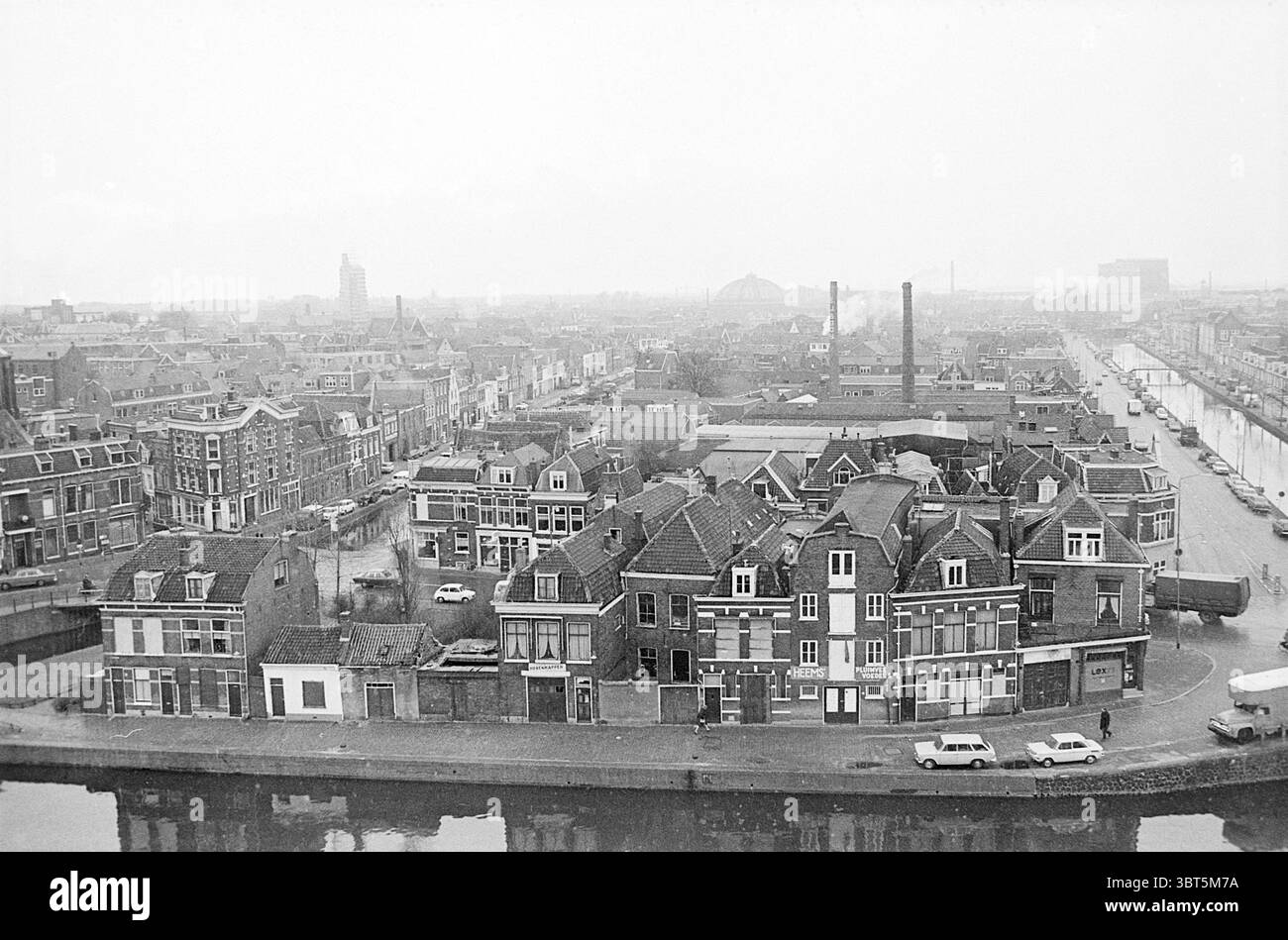 Übersichtsfoto von Burgwalbuurt Haarlem, Niederlande., Whizgle News, Dutch Desk, Niederlande, 1950 - 2000. Das Bild enthält diese Themen. Die Szene präsentiert eine weitläufige urbane Landschaft, die von dicht gepackten Gebäuden geprägt ist, von denen viele traditionelle Architekturstile mit Giebeldächern und komplizierten Fassaden zeigen. Die Strukturen bestehen hauptsächlich aus Ziegelsteinen, die verschiedene Grau-, Braun- und gedämpfte Farben zeigen, was der Umgebung ein Vintage-Flair verleiht. Im Vordergrund steht eine Ansammlung von Gebäuden in der Nähe des Wasserrandes, wo die ruhige Oberfläche t reflektiert Stockfoto