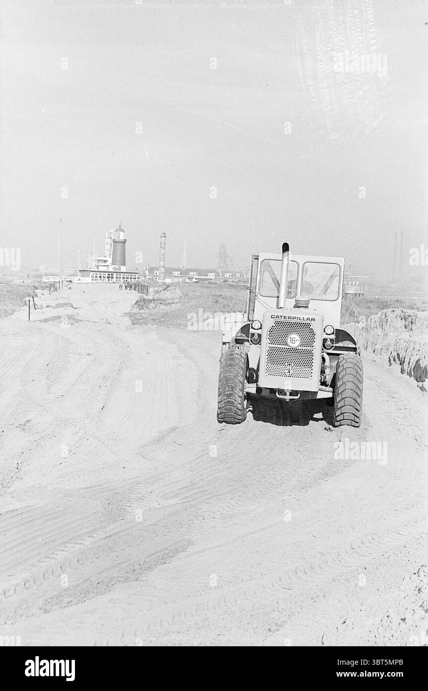 Tractor at Beach., Whizgle News, Dutch Desk, Niederlande, 1950 - 2000. Dies sind die Themen im Bild. Die Szene zeigt eine Industrielandschaft, die von schweren Maschinen und einem laufenden Bau- oder Aushubprozess geprägt ist. Ein großes, robustes Fahrzeug dominiert den Vorderboden, dessen markante Reifen das sandige Gelände erfassen. Das Fahrzeug ist in einer kräftigen, zweckmäßigen Farbe lackiert, mit einer auffälligen Zahl an der Seite, die sein funktionales Design unterstreicht. Im Hintergrund erheben sich verschiedene Strukturen vor einem trüben, bewölkten Himmel, was auf einen ausgedehnten Industriekomplex hindeutet. Diese b Stockfoto