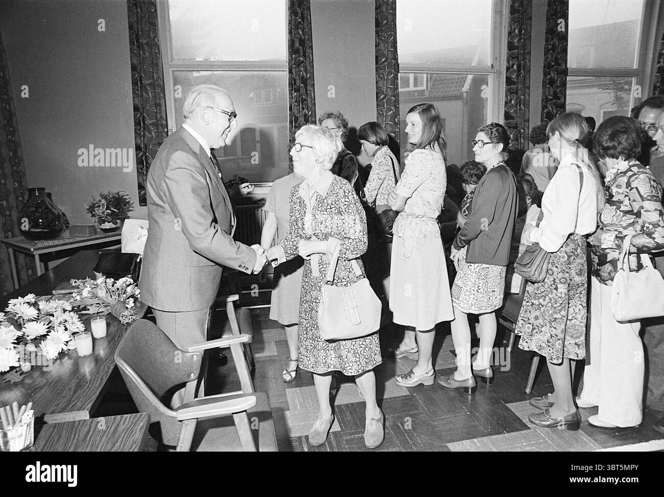 (Tagungs-)Raum, in dem ein älteres Paar geehrt wird, adressiert und gratuliert wird. Eine Menge. Whizgle News, Dutch Desk, Niederlande, 1950 - 2000. Dies sind die Themen im Bild. In einem geräumigen Innenbereich findet eine Zusammenkunft von Menschen statt, die von einer lebhaften Atmosphäre geprägt ist. Der Hintergrund ist mit großen Fenstern versehen, die weiches, diffuses Licht hereinlassen und ein Umgebungslicht erzeugen, das die Wärme der Szene erhöht. Eine Gruppe von Personen steht in der Schlange und wartet darauf, dass sie an der Reihe sind, um mit einer Person an der Spitze zu sprechen. Diese zentrale Figur, ein Mann in einem maßgeschneiderten Anzug, lächelt warm Stockfoto