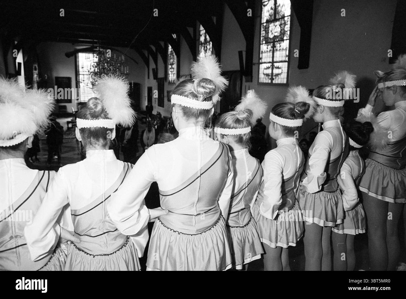 Carnival Key Offer, Whizgle News, Dutch Desk, Niederlande, 1950 - 2000 am 27-02-1992. Dies sind die Themen im Bild. In der Szene wird eine Gruppe von Darstellern in einer Probe oder einem Auftritt aufgenommen. Die Einzelpersonen sind in einem Halbkreis angeordnet, von hinten betrachtet, und zeigen ihre koordinierte Kleidung. Sie tragen passende Kostüme aus hellem Stoff, die mit Bändern und dekorativen Zierleisten verziert sind. Jedes Outfit wird durch einen unverwechselbaren Kopfschmuck mit flauschigen weißen Pompons ergänzt, die einen verspielten Kontrast zu ihren eleganten Frisuren bilden. Die Komposition zeigt die b Stockfoto