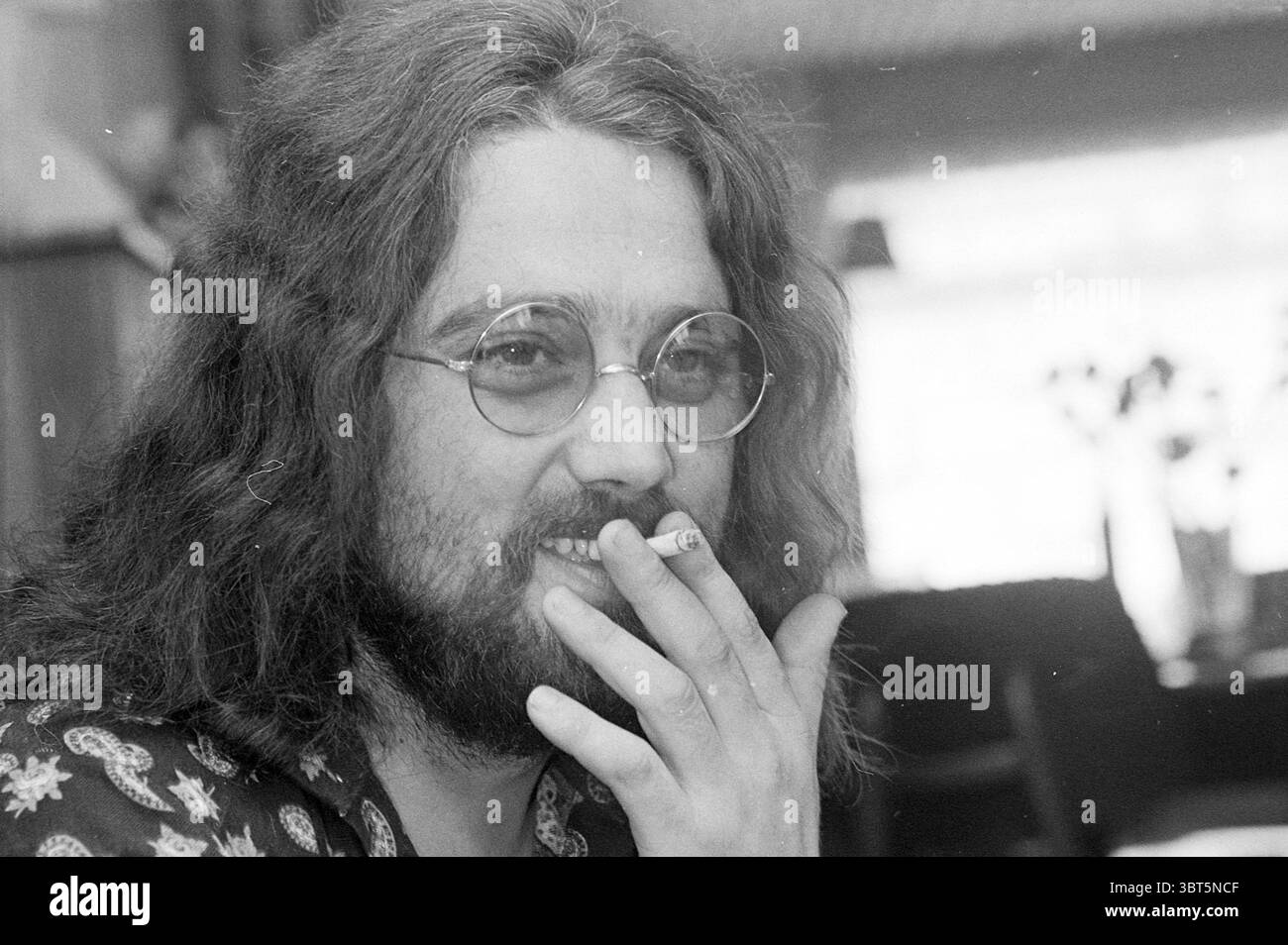 Porträt des Sängers Willem Duijn, bekannt als Big Mouth des Duos Mouth & MacNeal., Whizgle News, Dutch Desk, Niederlande, 1950 - 2000. Das Bild enthält diese Themen. In einem warm beleuchteten Innenbereich wird ein Mann mit langen, welligen, dunklen Haaren und einem Vollbart zentral in der Szene positioniert. Seine runde, metallgerahmte Brille verleiht ihm einen ausdrucksstarken Look, der ein Gefühl von Offenheit und Nachdenklichkeit widerspiegelt. Er hält eine Zigarette zwischen den Fingern und bringt eine lässige, aber kontemplative Atmosphäre in den Moment. Seine Kleidung ist mit einem gemusterten Hemd versehen, das auffällige, lebhafte Muster zeigt, die einen Hauch von hinzufügen Stockfoto