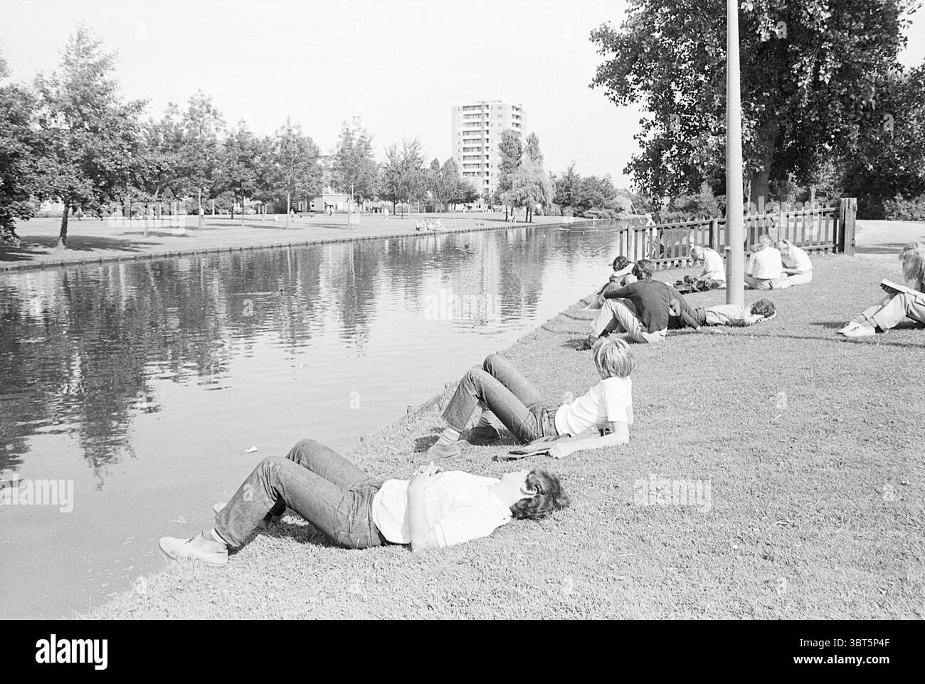 Menschen, die auf einem Kanal sonnen Haarlem Jan Gijzenkade Niederlande, Whizgle News, Dutch Desk, Niederlande, 1950 - 2000 am 08-1968. Diese Themen sind in der Abbildung dargestellt. In einer ruhigen Umgebung im Freien, ist ein ruhiger Wasserweg von grünem Gras gesäumt, der eine friedliche Atmosphäre schafft. Auf der linken Seite sitzt oder liegt eine Gruppe von Individuen auf dem Gras, die sich lässig am Ufer des Wassers befinden. Manche Menschen lehnen sich zurück, während ihre Köpfe zurückliegen, während andere tief in Gedanken oder Gesprächen zu einer entspannten Atmosphäre beitragen. Die Landschaft ist mit Reifen Bäumen bestückt, deren Blätter ein d Stockfoto