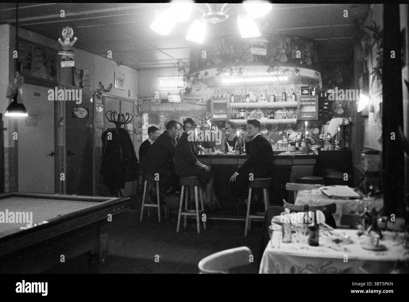 Family de Boer im Café Brabant Café, Whizgle News, Dutch Desk, Niederlande, 1950 - 2000 am 25-12-1960. Das Bild enthält diese Themen. Die Szene zeigt eine gemütliche, schwach beleuchtete Bar mit nostalgischer Atmosphäre. Im Vordergrund befindet sich ein großer Billardtisch auf einer Seite, dessen Grün sich leicht abgenutzt anfühlte, was darauf hindeutet, dass viele Spiele gespielt wurden. Die Wände sind mit verschiedenen Dekorationen verziert, die dem Raum Charakter und Wärme verleihen. An der Bar sitzt eine Gruppe von fünf Personen auf hohen Holzhockern, um sich zu unterhalten. Sie sind in Kleidung aus der Mitte des 20. Jahrhunderts gekleidet, was auf eine formelle oder halbförmige Form hindeutet Stockfoto