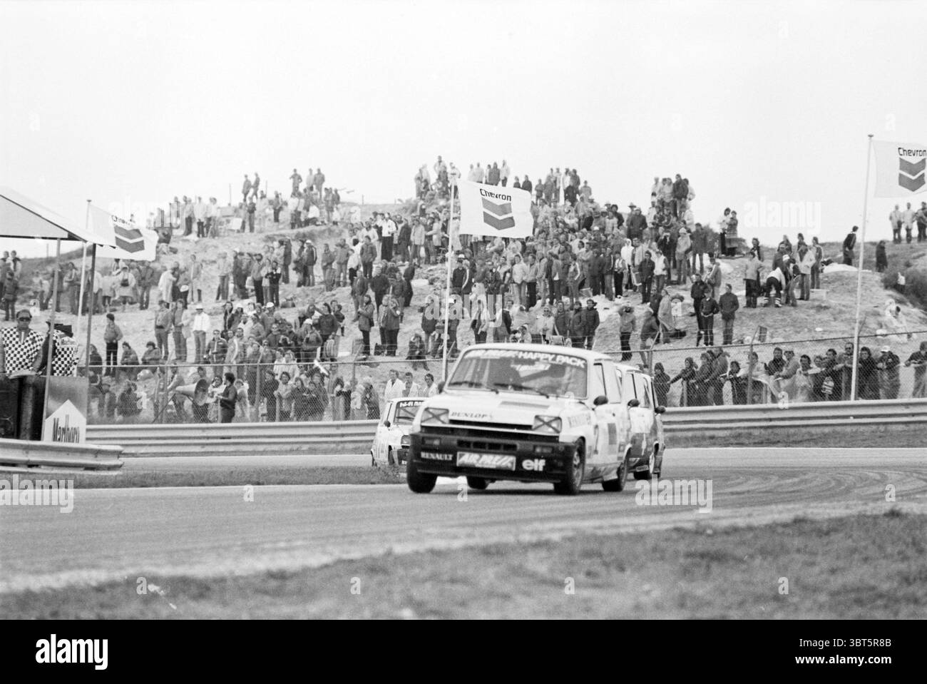 Formel I Abend + Fuego Renault, Whizgle News, Dutch Desk, Niederlande, 1950 - 2000 am 01-09-1980. Dies sind die Themen im Bild. Die Szene fängt die aufregende Atmosphäre eines Motorrennsports ein. Im Vordergrund zoomt ein klassischer Rennwagen, überwiegend weiß mit auffälligen Sponsorenlogos, um eine kurvenreiche Strecke. Das Design des Fahrzeugs mit seinem flachen Profil und seiner aerodynamischen Form weist auf Geschwindigkeit hin. Die Räder scheinen eine kleine Staubwolke hochzuschlagen, was die dynamische Action unterstreicht. Um die Strecke herum ist eine Menge Zuschauer zu sehen, die auf einem felsigen Hügel stehen. In einer Mischung gekleidet Stockfoto