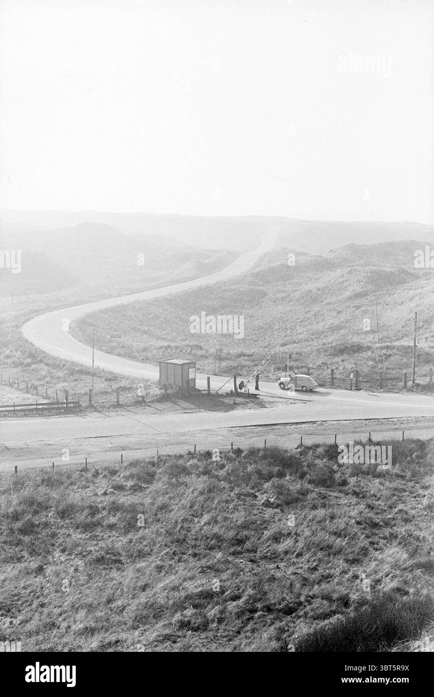 Barriere Velsen North - Beverwijk to the Beach Road Construction Road Planting, Whizgle News, Dutch Desk, Niederlande, 1950 - 2000 am 13-02-1964. Diese Themen sind in der Abbildung dargestellt. Die Szene fängt eine gewundene Straße ein, die sich anmutig durch eine weite, hügelige Landschaft schlängelt. Die mit grauem Asphalt gepflasterte Straße schlängelt sich in die Ferne und erzeugt ein Gefühl von Rätsel und Vorfreude auf das, was vor uns liegt. Das Gelände um die Straße herum ist geprägt von sanften Hügeln, bedeckt mit trockenem Gras und karger Vegetation, die gedämpft in Braun- und Grüntönen erscheinen. Auf einer Seite von t Stockfoto