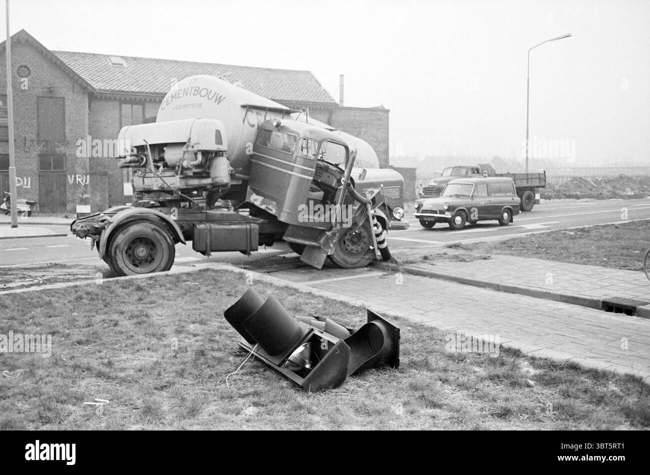 Auflieger Cementbouw, Whizgle News, Dutch Desk, Niederlande, 1950 - 2000 auf 12-1963. Dies sind die Themen im Bild. Die Szene zeigt eine etwas chaotische urbane Umgebung mit einer Mischung aus Fahrzeugen und industriellen Elementen. Im Vordergrund steht ein großer, klassischer Zementwagen, dessen abgerundete, sperrige Struktur ein Gefühl von Schwere vermittelt. Der mit einem Markennamen gekennzeichnete Tank des Staplers ist deutlich sichtbar, während das Fahrerhaus offenbar beschädigt wurde und sich unangenehm neigt, als ob es auf ein Hindernis gestoßen wäre. In der Nähe befindet sich ein älteres kleines Auto, vielleicht ein klassisches Modell, auf dem Ro Stockfoto