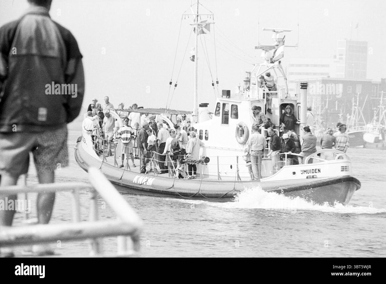 Tag der offenen Tür KNRM, Whizgle News, Dutch Desk, Niederlande, 1950 - 2000 am 05.05.1995. Das Bild enthält diese Themen. Die Szene fängt einen geschäftigen Moment auf dem Wasser ein, dominiert von einem Schiff, das sowohl praktisch als auch robust ist und eindeutig für den Seeverkehr konzipiert ist. Das Boot ist mit einer lebhaften Auswahl von Fahnen geschmückt, die im Wind flattern und auf Aktivität und Zweck hinweisen. Es ist von einem schäumenden Schwung umgeben, der sich hinter ihm erstreckt, während es sich durch das Wasser bewegt und ein Gefühl von Bewegung und Dringlichkeit vermittelt. An Bord ist eine vielfältige Gruppe von Personen sichtbar, von denen einige zusammen gruppiert sind, während andere es sind Stockfoto