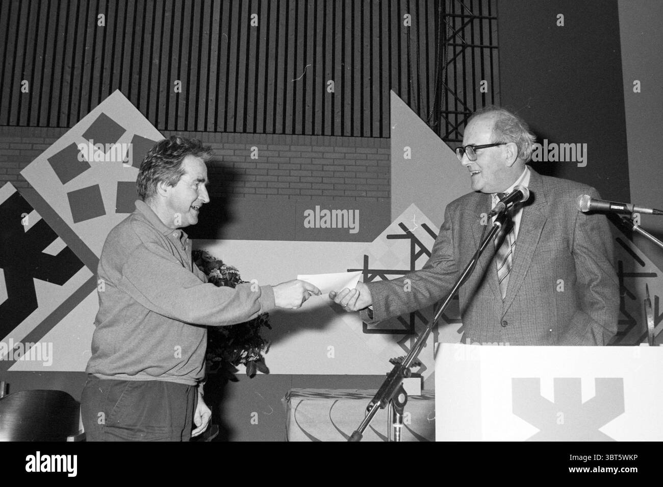 Verpflegung und Preisverteilung H.S.T. HST, Whizgle News, Dutch Desk, Niederlande, 1950 - 2000 am 28-01-1990. Das Bild enthält diese Themen. In der Szene sind zwei Männer an einem formellen Austausch beteiligt, der einen Moment der Anerkennung oder Auszeichnung vorschlägt. Ein Mann steht links, trägt ein dunkles Hemd und wirkt jugendlich und lebendig, mit kurzen, zerzausten Haaren. Er streckt seine Hand auf den zweiten Mann, der rechts steht. Der zweite Mann ist älter, in einem hellen Anzug und Krawatte gekleidet, mit einer Brille versehen, die ein Gefühl von Autorität und Weisheit vermittelt. Der Hintergrund ist mit einem großen abs ausgestattet Stockfoto