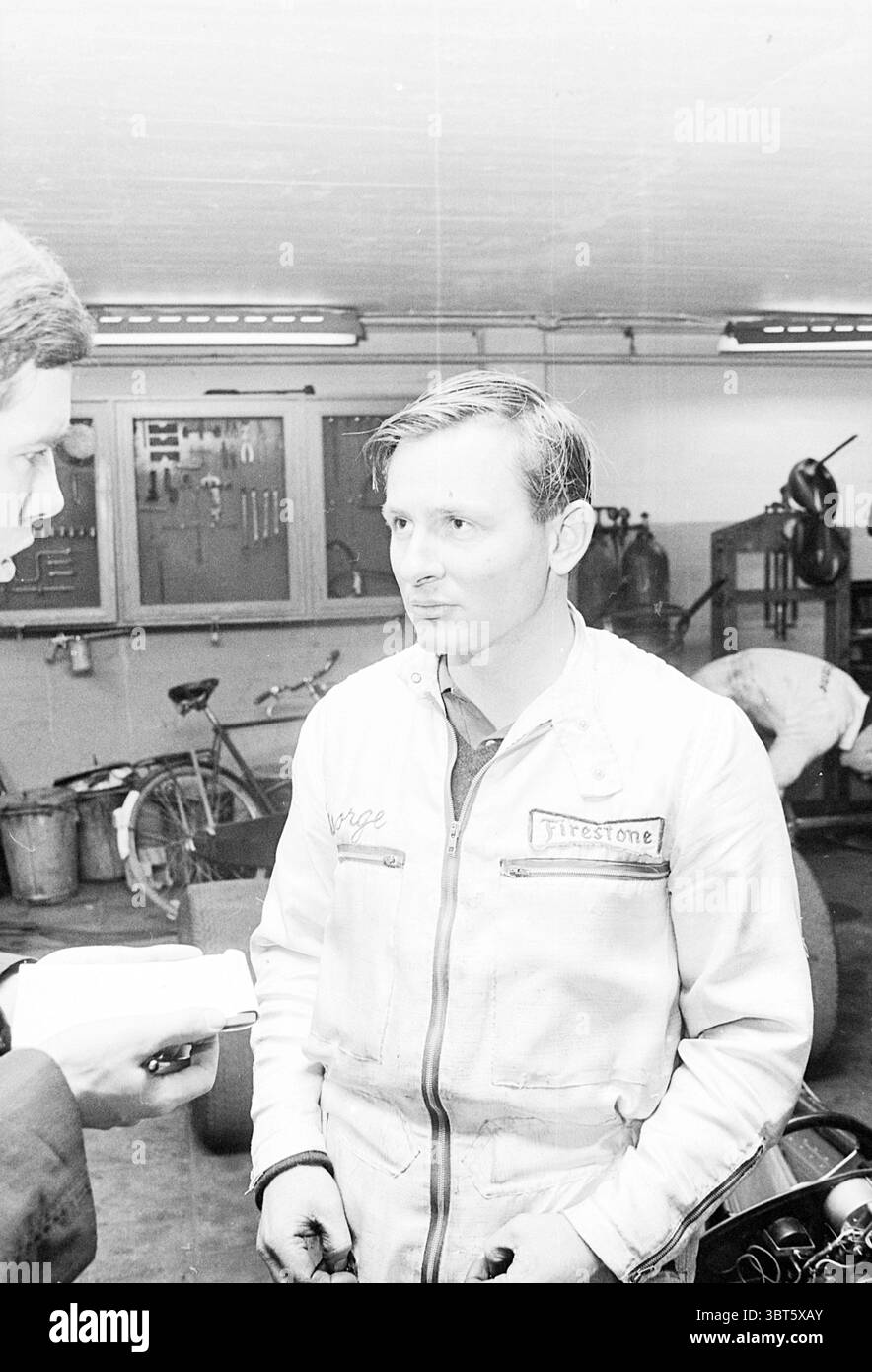 Bruce McLaren mit Race Zandvoort Persons, Whizgle News, Dutch Desk, Niederlande, 1950 - 2000 am 13.10.1965. Diese Themen werden in der Abbildung angezeigt. In einer schwach beleuchteten Garage steht ein junger Mann in der Mitte, gekleidet in einem hellen Mechaniker-Jumpsuit, der leicht faltig ist und subtile Gebrauchsspuren trägt. Der Jumpsuit ist mit einem Patch verziert, der möglicherweise auf einen Sponsor oder eine Marke hinweist, wodurch ein Gefühl der Authentizität für sein Handwerk entsteht. Sein Haar ist ordentlich zurückgekämmt, und sein Ausdruck wirkt besinnlich, als ob er in ein ernstes Gespräch verwickelt wäre. Links ist eine andere Person Stockfoto