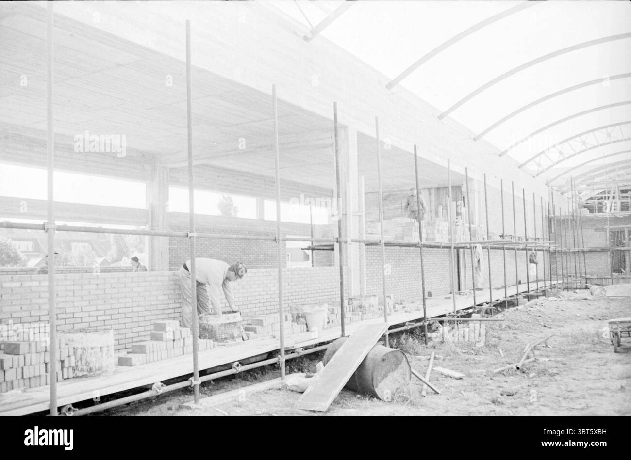 Industriehalle im Bau., Whizgle News, Dutch Desk, Niederlande, 1950 - 2000. Dies sind die Themen im Bild. Die Szene zeigt eine Baustelle in einem Fortschrittszustand. Langes, horizontales Gerüst verläuft an einer Seite und bietet Unterstützung für Arbeiter und Materialien. Ein Mann in Arbeitskleidung wird nach links positioniert, indem er vorsichtig Ziegelsteine an einer teilweise fertig gestellten Wand platziert. Seine Konzentration ist offensichtlich, als er einen Ziegel auf einer Kelle ausbalanciert. Die Wand ist sauber, einheitlich angeordnet von hellen Ziegeln, die einen Kontrast zu den rauen, erdigen Tönen der Umgebung bilden Stockfoto
