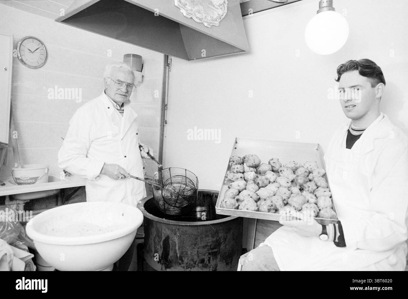 Mr. Brewer Oliebollenbäcker Oliebollen and Stalls, Whizgle News, Dutch Desk, Niederlande, 1950 - 2000 am 29-12-1989. Das Bild enthält diese Themen. In einer hell beleuchteten Küche werden zwei Personen hervorgehoben positioniert, die beide an der Zubereitung von Lebensmitteln beteiligt sind. Das Ambiente vermittelt eine Mischung aus Fleiß und Wärme, die durch das sanfte Leuchten der Deckenbeleuchtung unterstrichen wird. Auf der einen Seite steht ein älterer Mann mit weißem Haar, gekleidet mit einer weißen Schürze und Brille, neben einem Frittiergerät. Er konzentriert sich intensiv auf seine Aufgabe, ein Metallsieb zu halten, vielleicht um es wiederzufinden Stockfoto