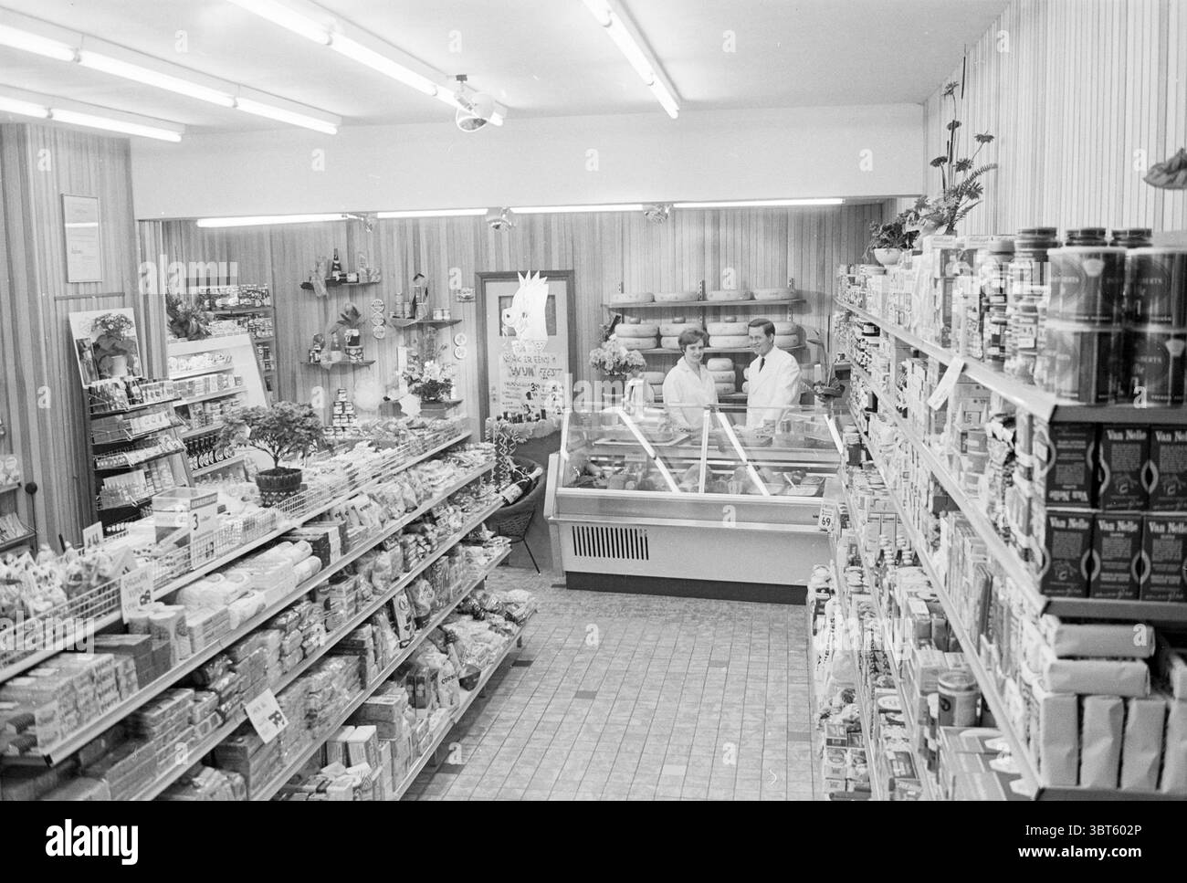 Warmerdam Interior, Whizgle News, Dutch Desk, Niederlande, 1950 - 2000 am 21-03-1968. Diese Themen werden in der Abbildung angezeigt. Die Szene zeigt ein nostalgisches Lebensmittelgeschäft mit einem einladenden Charme, der typisch für einen Markt aus der Mitte des 20. Jahrhunderts ist. Der Innenraum ist geräumig und gut beleuchtet, mit fluoreszierenden Deckenleuchten, die ein weiches, gleichmäßiges Leuchten über den Raum werfen. Die Holztäfelung an den Wänden hat einen warmen, erdigen Ton, der das Gefühl von Komfort und Vertrautheit verstärkt. Auf der linken Seite sind die Regale mit einer Vielzahl von Lebensmittelartikeln, einschließlich Konserven und Verpackungen, übersichtlich organisiert Stockfoto