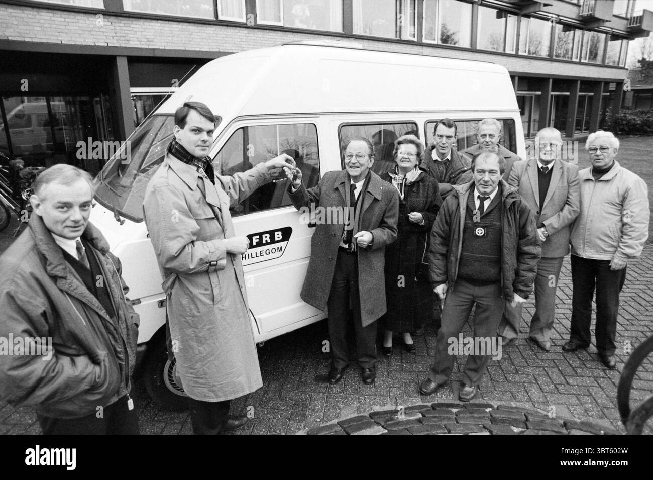 Transfer des Kleinbusses von FRB Hillegom Hillegom, Whizgle News, Dutch Desk, Niederlande, 1950 - 2000 am 15.12.1988. Dies sind die Elemente im Bild. In der Szene steht eine Gruppe von Individuen vor einem weißen Van, der in einem Bereich mit einem gepflasterten Boden geparkt ist. Der Van selbst hat ein schlichtes, klares Design und ein teilweise sichtbares Logo. Seine Farbe steht im Kontrast zu den gedämpften Tönen der Umgebung. Die Gruppe besteht aus acht Personen, überwiegend in Mänteln gekleidet, die für kühleres Wetter geeignet sind. Ihre Kleidung reicht von dunklen Tönen bis zu helleren Tönen, was eine Vielfalt von veranschaulicht Stockfoto