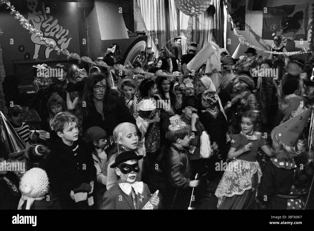 Carnival Velsen-Noord Carnival, Whizgle News, Dutch Desk, Niederlande, 1950 - 2000 am 10-02-1970. Dies sind die Themen im Bild. Die Szene ist voller Energie und Spannung, gefüllt mit einer großen Gruppe von Kindern, einige tragen bunte Kostüme und Masken, während sie an einer lebhaften Feier teilnehmen. Der Veranstaltungsort hat eine lebhafte Atmosphäre, geschmückt mit einer Mischung aus Dekorationen wie Luftschlangen und Ballons, die von der Decke und den Wänden hängen. Die Farben dominieren den Raum mit hellen Rot-, Gelb- und Blautönen, die sich vor einem dunkleren Hintergrund abheben. Kinder sind in den s verstreut Stockfoto