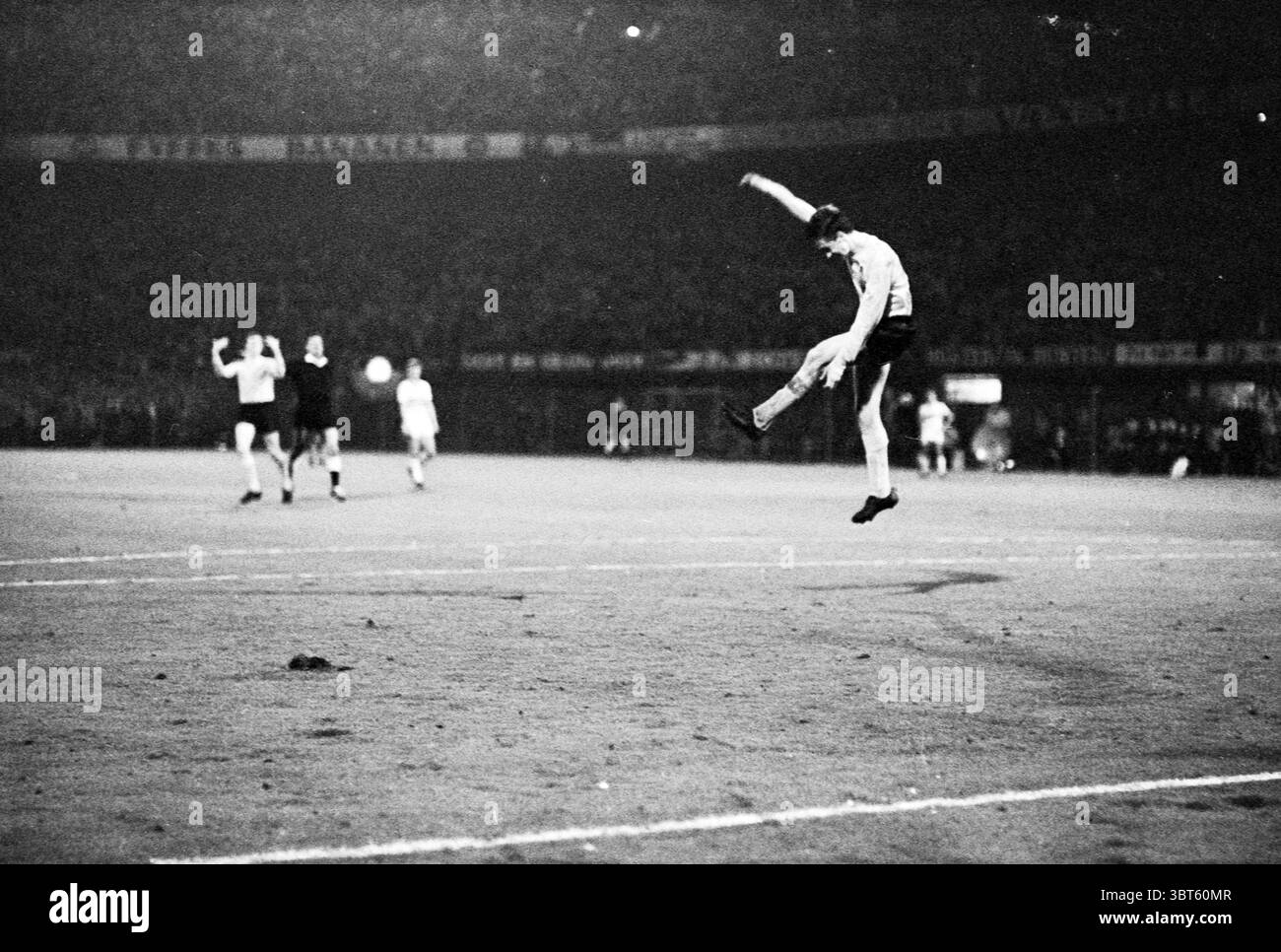 Fußballspiel in einem großen Stadion., Whizgle News, Dutch Desk, Niederlande, 1950 - 2000. Das Bild enthält diese Themen. Die Szene fängt einen dynamischen Moment auf einem Sportplatz während eines Spiels, wahrscheinlich eines Fußballspiels, mit einem Gefühl von Energie und Spannung ein. Im Vordergrund befindet sich ein Spieler mitten im Geschehen und hebt ein Bein hoch, was auf einen Versuch hindeutet, den Ball mit Kraft zu kicken. Seine Uniform ist dunkel und kontrastiert mit den helleren Farben anderer Spieler im Hintergrund. Die umliegenden Spieler zeigen verschiedene Haltungen, einige scheinen zu jubeln oder zu feiern, während andere sich auf die laufende pla konzentrieren Stockfoto