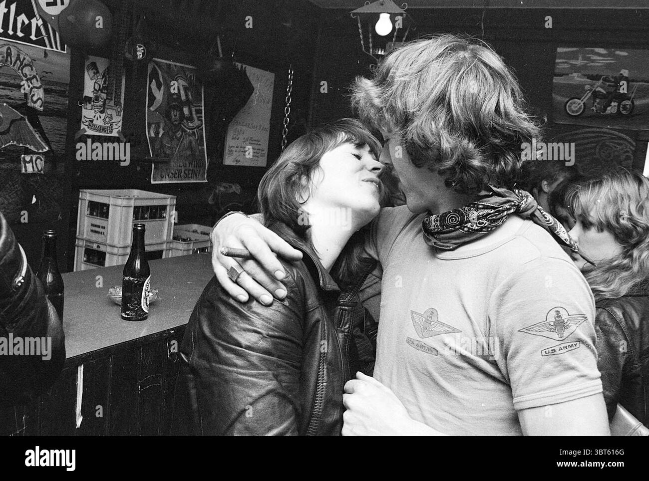 Hells Angels in Parklaan het Anker Hells Angels Motorradrennen Haarlem Parklaan Niederlande, Whizgle News, Dutch Desk, Niederlande, 1950 - 2000 am 13. 11. 1977. Dies sind die Themen im Bild. In einer lebendigen Umgebung stehen zwei Figuren im Vordergrund, die ein Gefühl von Intimität und Verbundenheit verkörpern. Das Paar ist nahe, ihre Köpfe berühren sich fast, während sie einen zärtlichen Moment teilen. Die Frau hat langes, dunkles Haar und trägt eine Lederjacke, die einen auffälligen Stil widerspiegelt. Ihr ausdrucksstarkes Verhalten deutet auf Freude und vielleicht einen Hauch von Verspieltheit hin. Der Mann mit zerzausten, mittellangen Haaren, Stockfoto