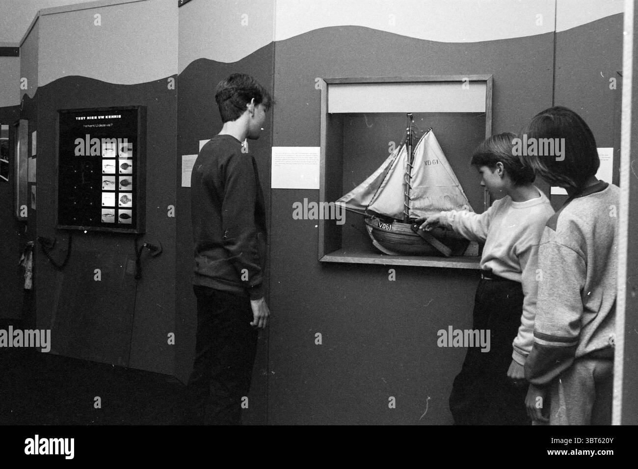 Fischereiausstellung P. Vermeulen Museum IJmuiden Ausstellungen Fischerei und andere Fischhändler Fischerei IJmuiden Niederlande, Whizgle News, Dutch Desk, Niederlande, 1950 - 2000 am 29-05-1984. Das Bild enthält diese Themen. In einer Galerie werden drei junge Menschen mit einer Ausstellung um ein Modellschiff beschäftigt. Das Modell ist aufwändig gestaltet und verfügt über weiße Segel, die sanft schweben, als ob sie von einer unsichtbaren Brise erfasst würden. Der in warmen Tönen lackierte Schiffsrumpf bildet einen Kontrast zum hellen weiß und verleiht dem Display Tiefe. Die Wände, die die Ausstellung umgeben, sind prädominiert Stockfoto