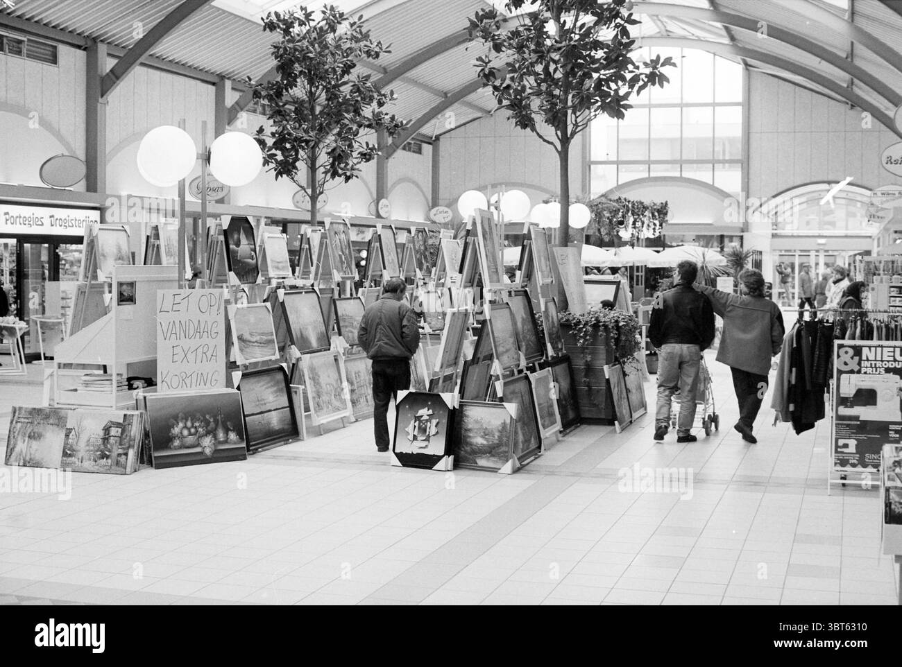 Malerverkauf Velserhof, Whizgle News, Dutch Desk, Niederlande, 1950 - 2000 am 28-01-1993. Diese Themen werden in der Abbildung angezeigt. Die Szene zeigt einen belebten Indoor-Markt oder Galerieraum, der durch eine lebhafte, aber entspannte Atmosphäre gekennzeichnet ist. Die Einstellung wird durch weiches, diffuses Licht beleuchtet, das durch eine hohe, bogenförmige Decke gefiltert wird. Die Architektur besticht durch eine Kombination aus Holz und modernen Materialien, die ein warmes, einladendes Gefühl verleihen. Im Vordergrund zeigen zahlreiche Staffeleien eine Vielzahl von Kunstwerken. Diese Stücke verfügen über eine reichhaltige Palette, die von tiefen, erdigen Tönen bis hin zu hellen, lebendigen K reicht Stockfoto