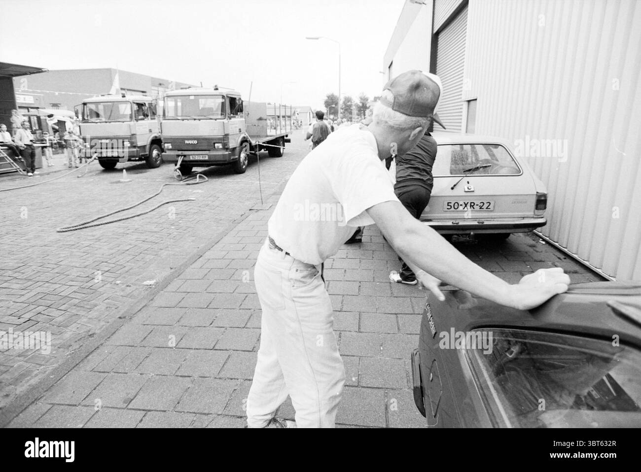 Tracker Pull Competition Haarlem the Netherlands, Whizgle News, Dutch Desk, Niederlande, 1950 - 2000 am 14.09.1991. Die Abbildung zeigt diese Themen. Die Szene zeigt ein geschäftiges Industriegebiet mit einer steinigen Straße, die von geparkten Fahrzeugen und Gebäuden gesäumt ist. Ein Mann, lässig gekleidet in einem T-Shirt und einer hellen Hose, lehnt sich an ein Auto, der Rücken wird dem Betrachter zugewandt. Er trägt eine Baseballmütze, die sein Gesicht leicht verdeckt, und ein sichtbares Bandana verleiht seinem Aussehen ein Element der Robustheit. Zu seiner Linken steht ein Oldtimer, dessen langweilige Töne auf das Alter hindeuten. In der Nähe zwei große trucks Stockfoto