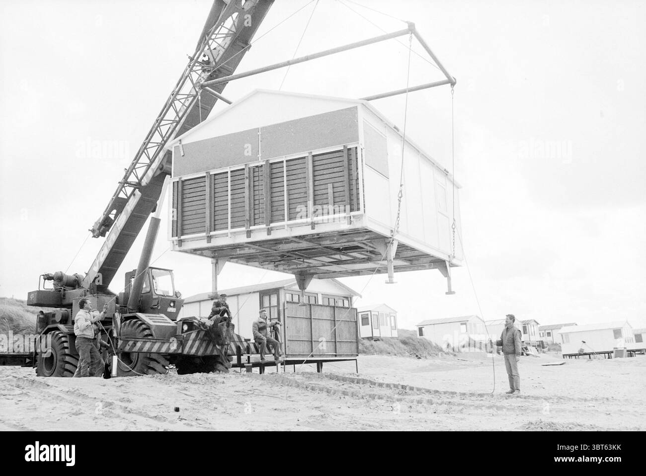 Abriss von Strandhäusern IJmuiden Beach and Beaches IJmuiden the Netherlands, Whizgle News, Dutch Desk, Niederlande, 1950 - 2000 am 24-09-1987. Das Bild enthält diese Themen. An der Küste ist ein großer Kran zu sehen, dessen Arm verlängert ist, um eine beträchtliche Struktur in die Luft zu heben. Die Szene ist monochrom und verleiht dem Moment ein nostalgisches oder historisches Gefühl. Das Gebäude, das angehoben wird, ähnelt einem kleinen Strandhaus, das sich durch ein schlichtes Design und eine Holzlattenfassade auszeichnet. Um den Kran herum sind mehrere Personen anwesend, von denen einige den Betrieb beobachten, während andere ihn beobachten Stockfoto