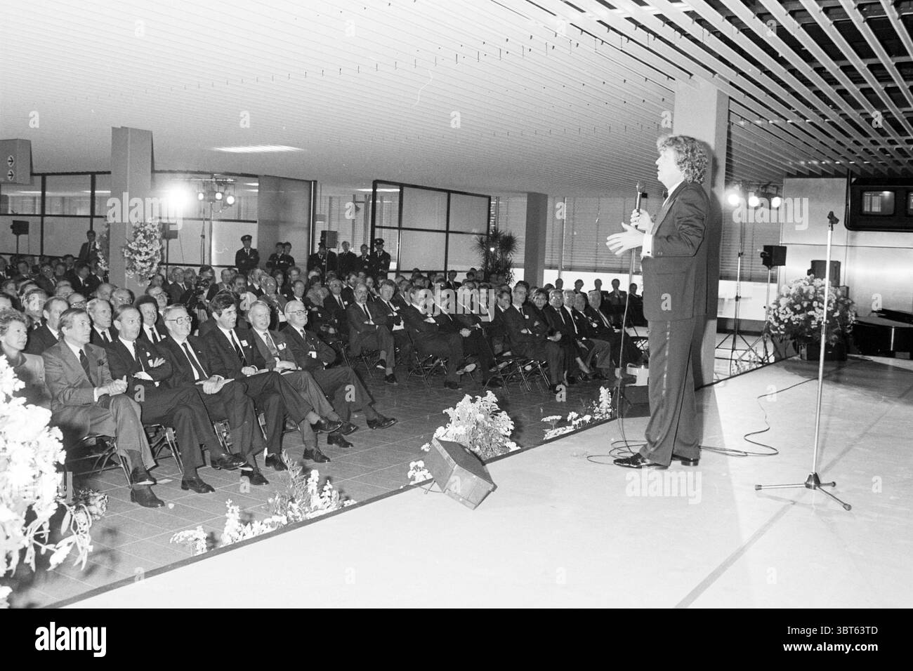 Eröffnung C-Pier Schiphol Schiphol, Whizgle News, Dutch Desk, Niederlande, 1950 - 2000 am 13.12.1987. Diese Themen sind in der Abbildung dargestellt. Die Szene verfügt über einen großen Auditorium mit einer Menge, die aufmerksam einem Lautsprecher auf einer erhöhten Bühne zugewandt ist. Der Redner, der in formeller Kleidung gekleidet ist, bewegt sich dynamisch, während er das Publikum anspricht. Die Menge besteht überwiegend aus Männern in Anzügen, wobei auch einige Frauen anwesend sind, die alle Ausdrucksformen von konzentrierter Betrachtung bis hin zu Engagement zeigen. Die Gesamtfarbpalette ist gedämpft und orientiert sich an monochromatischen Farbtönen mit Schwarz-, Grau-, Stockfoto