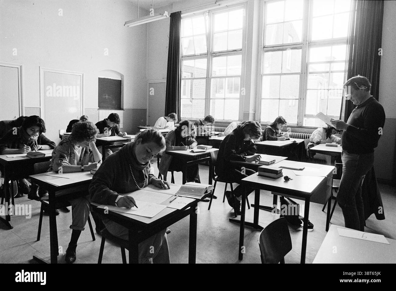 Abschlussprüfung Gymnasium Velsen Zuid Exam Velsen-Zuid, Whizgle News, Dutch Desk, Niederlande, 1950 - 2000 am 26-04-1985. Dies sind die Themen im Bild. Die Szene spielt in einem Klassenzimmer mit natürlichem Licht, das durch große Fenster hereinströmt und den Raum mit einem sanften Leuchten beleuchtet. Die Wände sind in einem hellen Farbton gestrichen, was die luftige Atmosphäre des Zimmers unterstreicht. Die Schreibtische sind in Reihen angeordnet, die jeweils von Schülern besetzt sind, die sich über ihre Arbeit gebeugt und in ihre Aufgaben eingelassen haben. Ihre Ausdrücke variieren; einige sehen konzentriert aus, während andere tief in Gedanken erscheinen. Jeder Schüler ist mit Schreiben m ausgestattet Stockfoto