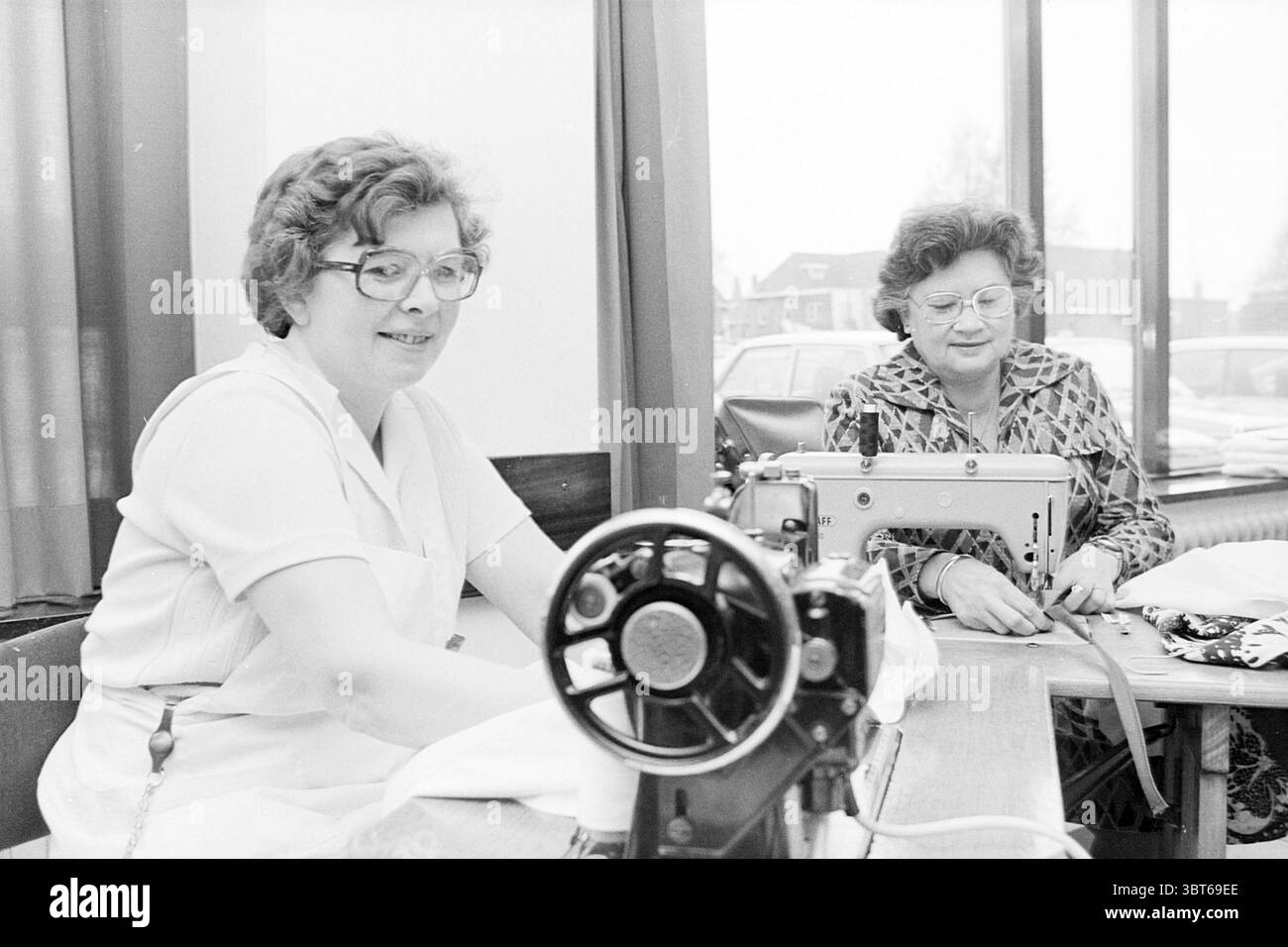 2 Damen aus der Leinen Abteilung Zeeweg Krankenhaus IJmuiden Zeeweg Niederlande, Whizgle News, Dutch Desk, Niederlande, 1950 - 2000 am 04-04-1977. Diese Themen werden in der Abbildung angezeigt. Die Szene zeigt zwei Frauen, die am Nähen arbeiten, an einem langen Tisch sitzen, der sich über einen gut beleuchteten Innenraum erstreckt. Eine Frau mit lockigem Haar und Brille lächelt, während sie eine Nähmaschine bedient. Der metallische Mechanismus leuchtet im hellen Licht. Sie trägt ein knackiges weißes Outfit, das einen Kontrast zu dem reichen, dunklen Holz des Tisches unter ihr bildet. Die zweite Frau, etwas älter, trägt ein gemustertes Kleid Stockfoto