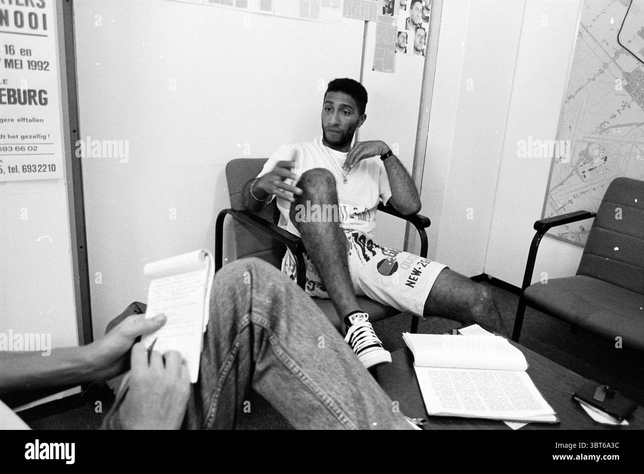 Lesley Hellings Fußballtrainer beim FC de Sloterplas und den Stormvogels. Amsterdam Niederlande, Whizgle News, Dutch Desk, Niederlande, 1950 - 2000 am 07-07-1992. Diese Themen werden in der Abbildung angezeigt. In einem bescheidenen Innenbereich sitzt ein Mann entspannt auf einem modernen Stuhl, der leicht außermittig positioniert ist. Er trägt ein lässiges Outfit, zu dem ein weißes T-Shirt mit Grafiken und knielange Shorts gehören. Seine Beine sind überkreuzt und zeigen ein Paar gemusterte Socken und klassische Sneakers. Seine Haltung ist entspannt und doch selbstbewusst, mit einem Arm auf der Armlehne, während er mit dem anderen Gesten, Su, spielt Stockfoto