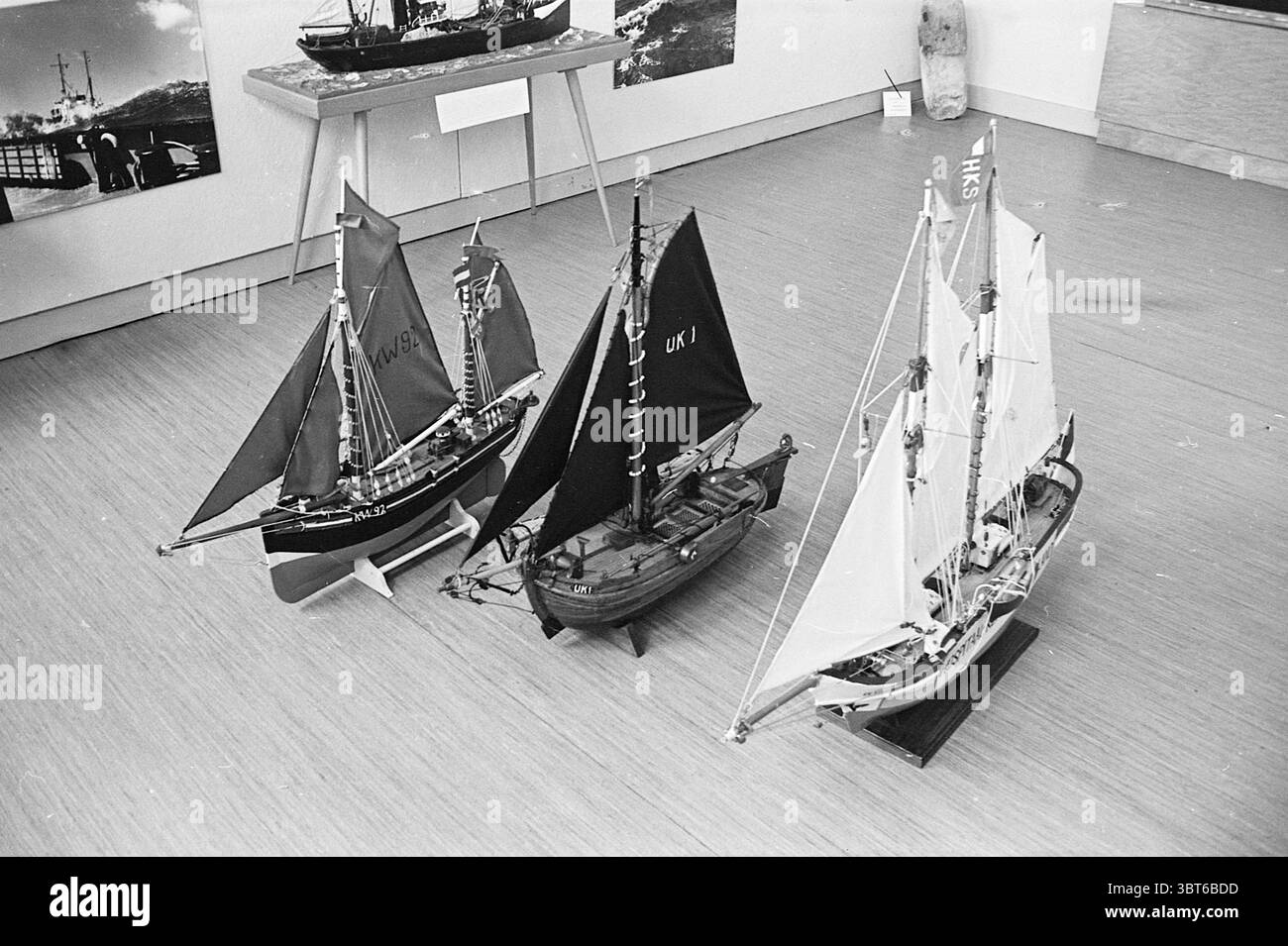 3 Schiffsmodelle Pieter Vermeulen Museum Ships, Whizgle News, Dutch Desk, Niederlande, 1950 - 2000 am 04-08-1964. Die Abbildung zeigt diese Themen. Die Szene zeigt drei aufwändig gefertigte Modellboote auf einem glatten, hellen Holzboden. Die Modelle unterscheiden sich in Design und Größe und weisen jeweils einzigartige Merkmale auf. Auf der linken Seite hebt sich ein Modell mit dunklen Segeln hervor, dessen rote und schwarze Farben einen auffälligen Kontrast zur helleren Umgebung bilden. Er verfügt über einen detaillierten Rumpf, komplett mit sichtbarer Takelage und Miniatur-Hardware, die ein Gefühl von Authentizität widerspiegelt. In der Mitte, A Stockfoto