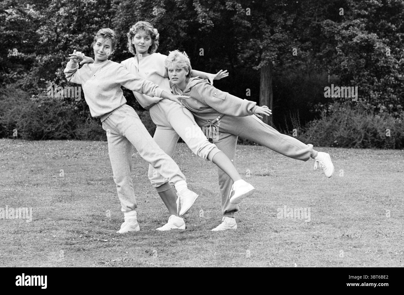 Pony Aerobic Tanzbekleidung + Schuhe Pony Sports, Whizgle News, Dutch Desk, Niederlande, 1950 - 2000 am 29-06-1983. Diese Themen werden in der Abbildung angezeigt. In dieser Szene interagieren drei Personen spielerisch vor einem Hintergrund von üppigem Grün. Die Umgebung ist ein weitläufiges Grasgebiet, umgeben von dichtem Laub, das der Umgebung einen erdigen Ton verleiht. Die drei Personen tragen dazu passende Sweatsuits, überwiegend in hellgrauen und weichen Pastelltönen, die die natürliche Umgebung ergänzen. Ihre Outfits versprühen eine lässige, entspannte Atmosphäre, die einen Tag voller Freizeit oder Aktivität anregt. Jeder Perso Stockfoto