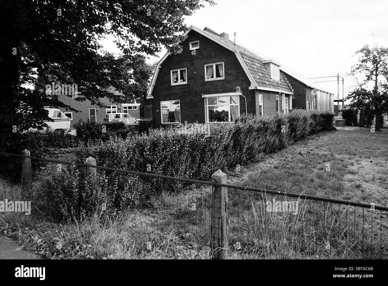 Schipholweg Farm Badhoevedorp, Whizgle News, Dutch Desk, Niederlande, 1950 - 2000 am 07-08-1990. Diese Themen sind in der Abbildung dargestellt. Die Szene zeigt ein malerisches, zweistöckiges Haus mit einem unverwechselbaren A-Rahmen Dach, das durch seine dunkle Ziegelfassade gekennzeichnet ist. Das Gebäude liegt etwas von der Straße entfernt, umgeben von einer gut gepflegten Hecke, die eine klare Grenze für das Anwesen schafft. Grünes Gras bedeckt den Hof teilweise, was auf das jüngste Wachstum hindeutet, während trockene Grasflecken darauf hindeuten, dass es in anderen Bereichen nicht regnet. Auf einer Seite des Hauses ist die Umrisse einer Garage oder eines Versorgungsraums zu sehen Stockfoto