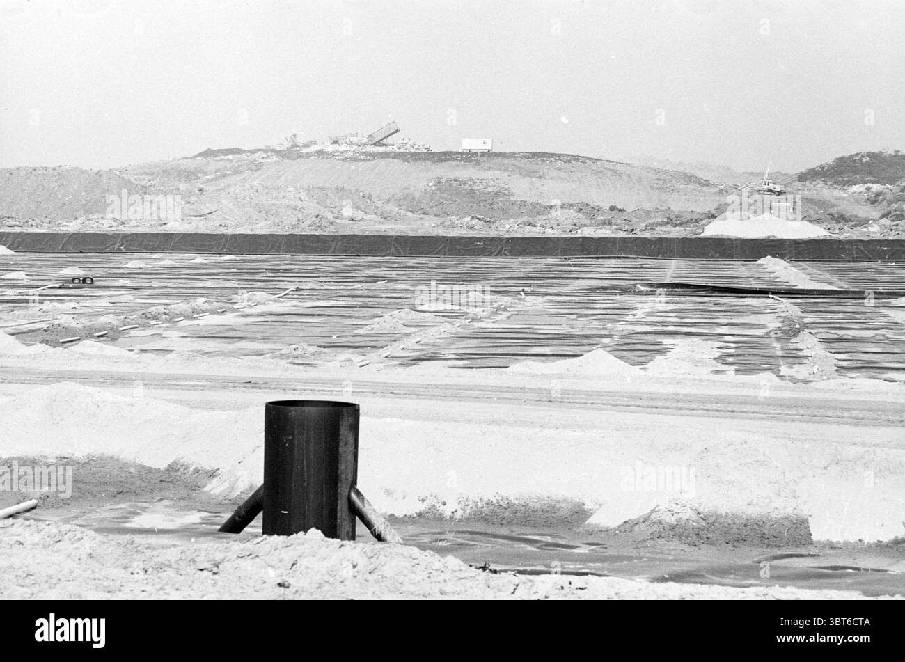 Oostbroekerweg neues Gelände durch Tunnelterrains Tunnel Velsen-Zuid Oostbroekerweg, Whizgle News, Dutch Desk, Niederlande, 1950 - 2000 am 08-09-1989. Dies sind die Elemente im Bild. Die Szene zeigt eine ausgedehnte Industrielandschaft, gekennzeichnet durch große Flächen von hellem Sandmaterial, möglicherweise Salz oder einem anderen Mineral, das sich in organisierten Reihen über den Boden verteilt. Im Vordergrund erhebt sich eine dicke, dunkle zylindrische Struktur aus dem Boden, die sich stark von den hellen Farbtönen der Umgebung abhebt. Der Boden ist flach und scheint sich endlos zu dehnen, mit Flecken o Stockfoto