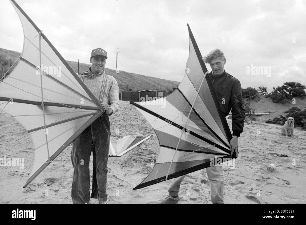 Skating/Kite Zandvoort Zandvoort, Whizgle News, Dutch Desk, Niederlande, 1950 - 2000 am 20-09-1990. Dies sind die Themen im Bild. Die Szene zeigt zwei Personen, die an einem Sandstrand stehen und jeweils einen farbenfrohen Drachen mit auffälligen geometrischen Mustern halten. Die Drachen, die in leuchtenden Farben wie Rot, Blau, weiß und Schwarz gebaut sind, sind so konzipiert, dass sie die Aufmerksamkeit vor dem gedämpften, bewölkten Himmel erregen. Die beiden Figuren sind lässig gekleidet; die eine trägt einen hellen Pullover und eine Mütze, die andere trägt eine dunkle Jacke mit Reißverschluss. Beide haben ein ausdrucksstarkes Lächeln, das ein Gefühl von Freude und Anti vermittelt Stockfoto
