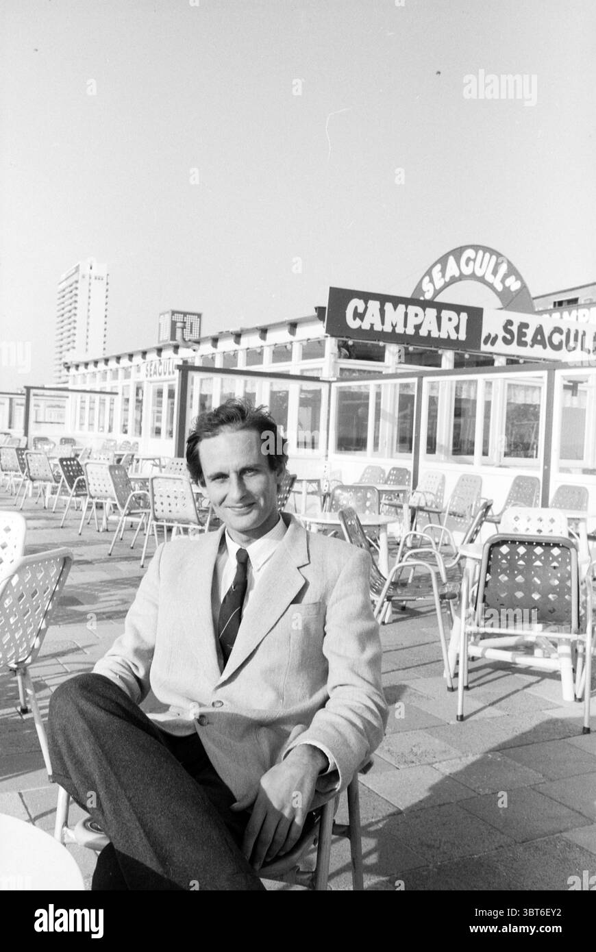 Herr Koopman über Zandvoort Beach Persons Zandvoort, Whizgle News, Dutch Desk, Niederlande, 1950 - 2000 am 04-05-1988. Die Abbildung zeigt diese Themen. Am Meer sitzt ein Mann zwanglos in einem Stuhl und strahlt ein entspanntes und selbstbewusstes Verhalten aus. Er trägt eine helle Anzugsjacke über einer dunklen Krawatte, was zu einem stylischen, aber dennoch zugänglichen Look beiträgt. Sein Haar ist ordentlich gekämmt, was seinem Aussehen einen Hauch von Raffinesse verleiht. Um ihn herum befinden sich zahlreiche leere Stühle, die in ordentlichen Reihen auf einer gepflasterten Terrasse angeordnet sind, die auf einen beliebten Treffpunkt hinweisen. Die Stühle mit ihren markanten perforierten Stühlen Stockfoto