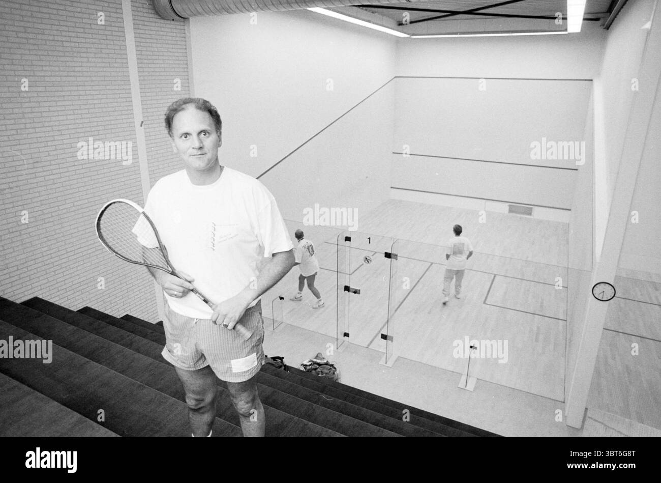 Erik Hooijman Squash Hoofddorp the Netherlands, Whizgle News, Dutch Desk, Niederlande, 1950 - 2000 am 16-09-1991. Das Bild enthält diese Themen. In einem geräumigen Squashplatz, der durch natürliche und künstliche Beleuchtung gut beleuchtet ist, steht ein Mann prominent auf einer Treppe, die zum Platz führt. Er hält einen Squashschläger in der rechten Hand, trägt ein lässiges weißes T-Shirt und hellgraue Shorts, die auf Komfort und Bewegung hinweisen. Sein Ausdruck ist freundlich, mit einem selbstbewussten Lächeln, das zeigt, dass er bereit ist, sich am Spiel zu beteiligen. Der Platz selbst verfügt über einen polierten Holzboden, der reflektiert Stockfoto