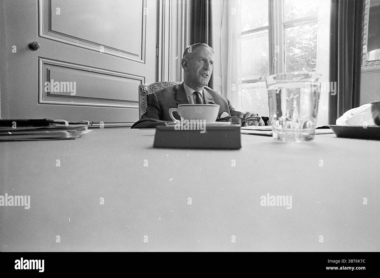 Porträt Mr. Van Dokkum Porträts, Whizgle News, Dutch Desk, Niederlande, 1950 - 2000 am 06-09-1971. Das Bild enthält diese Themen. Die Szene zeigt einen Mann, der an einem Tisch sitzt und sich in Gesprächen befindet. Er ist formell gekleidet, trägt einen Anzug, und sein Ausdruck vermittelt ein Gefühl der Nachdenklichkeit. Die Einrichtung scheint ein elegant eingerichteter Raum mit klassischem Dekor zu sein, in dem große Fenster natürliches Licht einlassen und der Umgebung ein warmes Licht verleihen. Auf dem Tisch befindet sich eine einfache weiße Tasse, die sich für Tee oder Kaffee eignet, neben einem klaren Glas Wasser, das das Licht wunderbar reflektiert. In der Zukunft Stockfoto