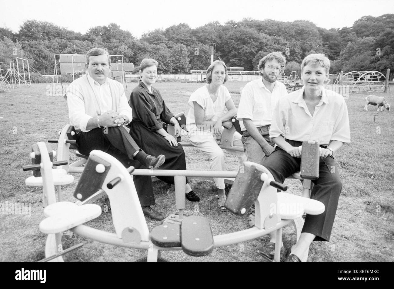Steigen Sie auf dem neuen Spielplatz IJmuiden Games and Toys IJmuiden the Netherlands, Whizgle News, Dutch Desk, Niederlande, 1950 - 2000 am 22-08-1989. Dies sind die Themen im Bild. In einer ruhigen Umgebung im Freien versammelt sich eine Gruppe von fünf Erwachsenen auf spielerisch gestalteten Schaukelpferden in Kindergröße. Die Szene ist in sanftes, diffuses Licht getaucht, das typisch für einen bewölkten Tag ist, was dem Treffen eine sanfte, dezente Stimmung verleiht. Die Einzelpersonen tragen legere Kleidung. Auf der einen Seite trägt ein Mann mit Schnurrbart ein weißes Hemd, das ein Gefühl der Leichtigkeit vermittelt. Neben ihm eine Frau in einem schwarzen Kleid Stockfoto