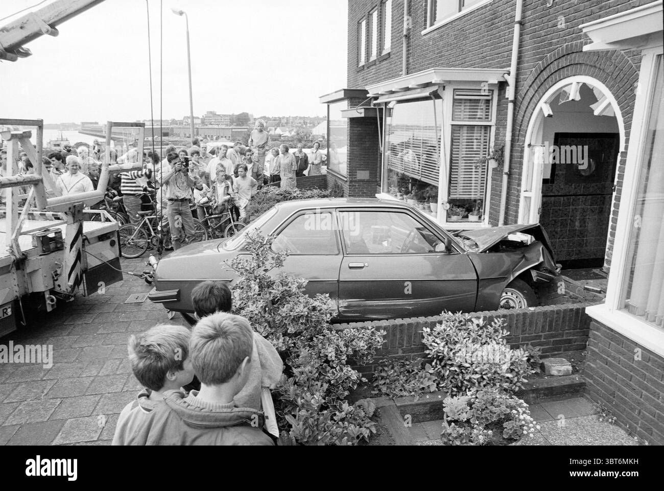 Auto trifft die Haustür der Linnaeusstraat 3 IJmuiden IJmuiden Linnaeusstraat Niederlande, Whizgle News, Dutch Desk, Niederlande, 1950 - 2000 am 11-06-1990. Diese Themen sind in der Abbildung dargestellt. In einer belebten Stadtszene ist ein Auto gegen die Seite eines Backsteinhauses gestürzt, was einen auffälligen Blickpunkt bildet. Das kompakte Fahrzeug befindet sich in einem ungünstigen Winkel und weist erhebliche Schäden am vorderen Ende auf, was auf die Kraft des Aufpralls hindeutet. Das Äußere des Hauses hat ein klassisches Design mit großen Fenstern, und der Bereich um den Eingang ist mit kleinen Sträuchern und Dekoraten geschmückt Stockfoto