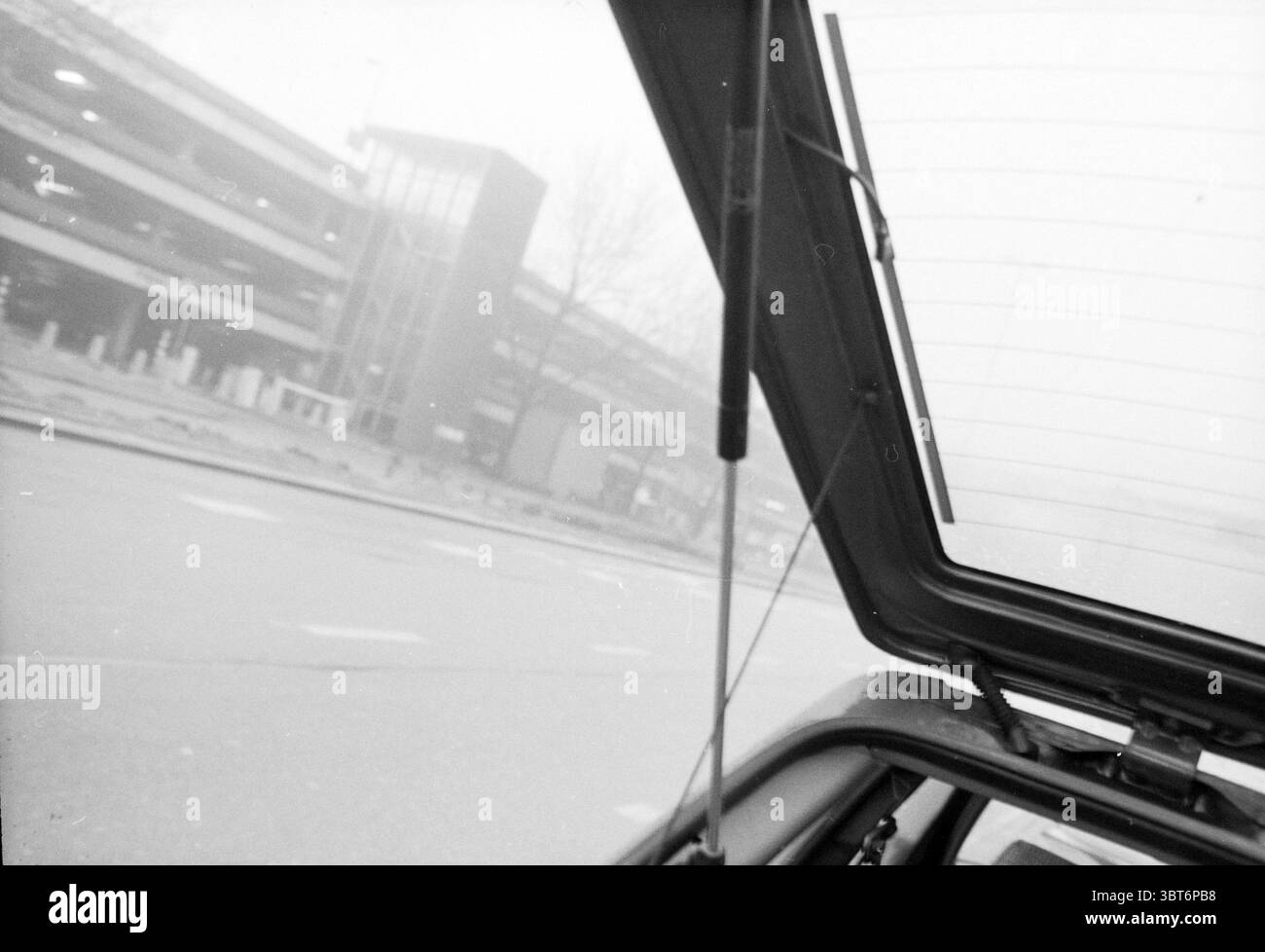 Heckklappe eines Autos im Nebel, Whizgle News, Dutch Desk, Niederlande, 1950 - 2000. Diese Themen werden in der Abbildung angezeigt. Die Szene präsentiert eine monochromatische Ansicht, die von Grau- und Weißtönen dominiert wird und ein Gefühl von düsterer Ruhe weckt. Im Vordergrund zeigt ein teilweise geöffneter Kofferraum einen Blick auf das Innere des Fahrzeugs. Die scharfen Winkel des Metalls bilden eine kontrastierende Linie gegen den weichen Nebel. Der Kofferraumdeckel ist offen und verleiht dem ansonsten ruhigen Ambiente einen Hauch von Aktivität. In der Mitte erscheint ein Parkhaus, obwohl es in Nebel gehüllt ist, was seine Details und Kontinuität verringert Stockfoto
