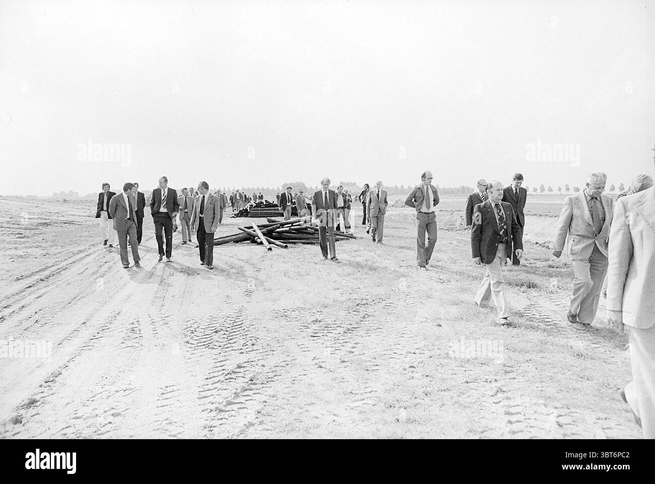 Durchschn. Besuche Empfehlen Sie Haarlemmermeer an recr. Plan H'dorp Haarlemmermeer Municipal Council, Whizgle News, Dutch Desk, Niederlande, 1950 - 2000 am 19.09.1975. Diese Themen sind in der Abbildung dargestellt. Die Szene zeigt eine Gruppe von Individuen, die auf einem unbefestigten Pfad laufen, der sich bis in die Ferne erstreckt. Der Boden ist mit Reifenspuren strukturiert, die auf frühere Fahrzeuge hinweisen, die den Bereich durchquert haben. Um den Weg herum ist die Landschaft meist flach, mit kargem Gras geschmückt, was zu einem etwas kargen Aussehen beiträgt. Im Hintergrund befinden sich Baumreihen, die eine verschwommene Linie gegen Th bilden Stockfoto