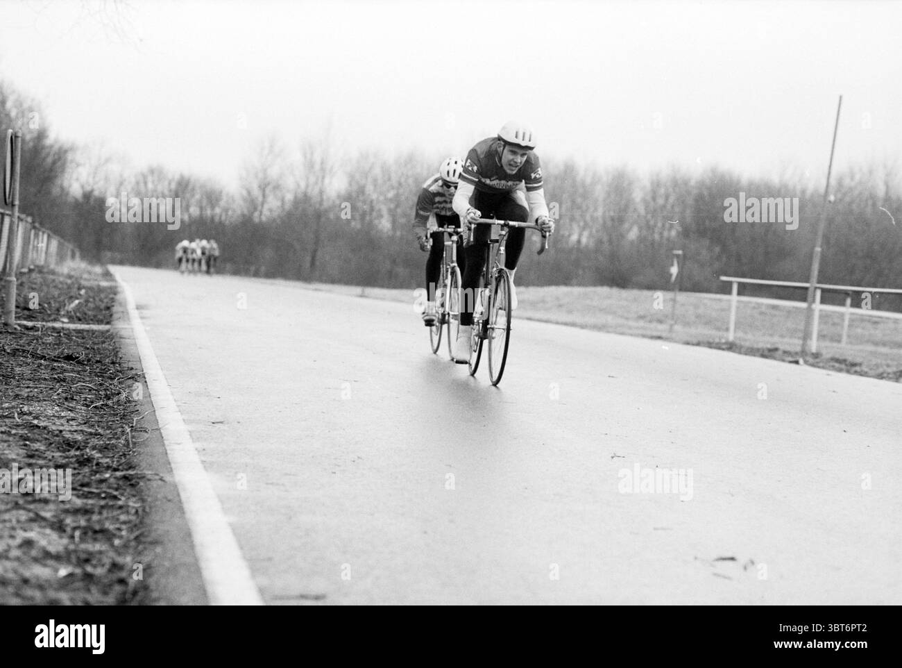 Radfahren Spaarnwoude Spaarnwoude, Whizgle News, Dutch Desk, Niederlande, 1950 - 2000 am 18-12-1993. Diese Themen sind in der Abbildung dargestellt. Die Szene zeigt ein dynamisches Radrennen auf einer breiten, glatten Asphaltstraße. Der Pfad verläuft durch eine leicht feuchte Umgebung, die eine gedämpfte Palette von Grau- und Brauntönen aufgrund des jüngsten Regens reflektiert. Im Vordergrund dominieren zwei Radfahrer die Komposition, deren intensive Ausdrücke und sportliche Haltungen ein Gefühl von Wettbewerb und Entschlossenheit vermitteln. Beide Fahrer tragen passgenaue Radtrikots in verschiedenen Farben – Dunkelblau und Rot im Kontrast zu A Stockfoto