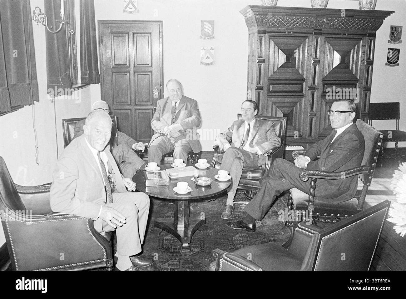 1. Arbeitstag Burg. De Gou Mayors, Whizgle News, Dutch Desk, Niederlande, 1950 - 2000 am 01-07-1969. Dies sind die Elemente im Bild. In einem freundlich eingerichteten Zimmer sitzen fünf Männer um einen eleganten runden Tisch. Der Tisch ist mit kleinen weißen Tassen und Untertassen gedeckt, was auf einen Moment der sozialen Bindung hindeutet, vielleicht bei Kaffee oder Tee. Die Wände sind mit dunkler Holztäfelung verziert, und darüber deuten subtile historische Motive auf eine formelle Atmosphäre hin. Der Raum verfügt über große Fenster, die weiches, diffuses Licht hereinlassen und eine sanfte Atmosphäre schaffen. Das Licht sorgt für ein sanftes Leuchten, das den Reichtum verstärkt Stockfoto