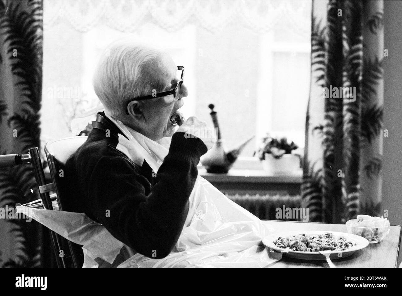 Bericht Janskliniek Nursing Homes Haarlem Ridderstraat Niederlande, Whizgle News, Dutch Desk, Niederlande, 1950 - 2000 am 24-03-1988. Das Bild enthält diese Themen. Die Szene zeigt einen älteren Mann, der an einem Esstisch sitzt und sich mit dem Essen beschäftigt. Er trägt dunkle Kleidung, ergänzt durch einen hellen Kragen und trägt eine Brille, die seinen konzentrierten Ausdruck unterstreicht. Eine Kunststoffschürze hängt über ihn und betont den praktischen Aspekt der Umgebung, während er seine Mahlzeit genießt. Der Tisch ist mit einem Teller geschmückt, der wie eine herzhafte Mahlzeit aussieht, umgeben von bescheidenen Utensen Stockfoto