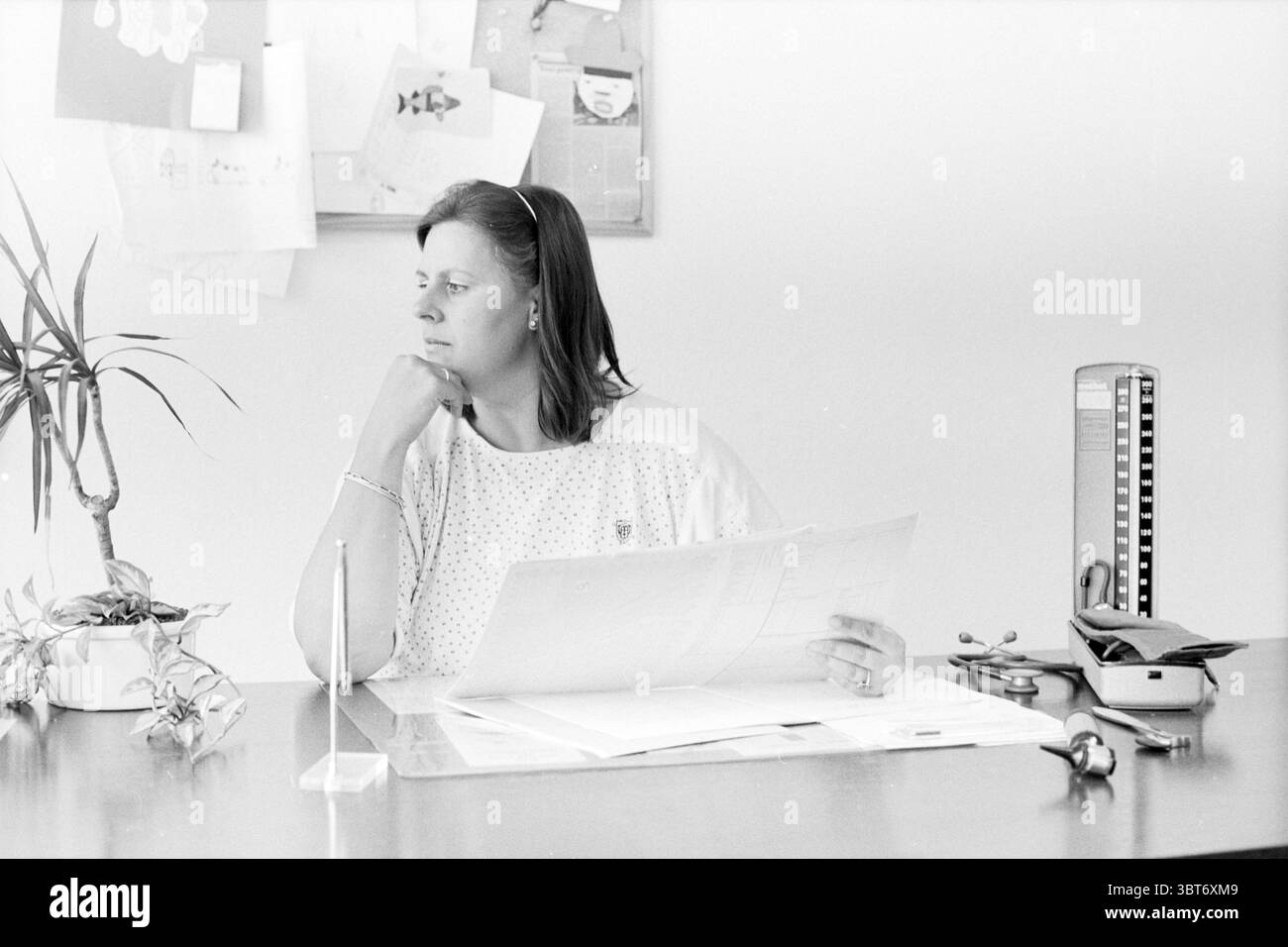 Frau Eerdenburg Schularzt Hillegom Ärzte Hillegom, Whizgle News, Dutch Desk, Niederlande, 1950 - 2000 am 14-07-1988. Die Abbildung zeigt diese Themen. In einem leise beleuchteten Bürobereich sitzt eine Frau an einem Schreibtisch und strahlt einen Hauch von Besinnung aus. Ihr nachdenklicher Ausdruck deutet darauf hin, dass sie tief in der Analyse ist, wobei ihr Kinn auf einer Hand ruht. Sie trägt ein helles Kleid, das sich sanft über ihre Schultern zieht und sich subtil mit den dunkleren Tönen des hölzernen Schreibtisches abhebt. Der Schreibtisch selbst ist mit verschiedenen Gegenständen geschmückt: Einer Topfpflanze mit schmalen Blättern, die einen Hauch von Natur verleiht, und einem Büro Stockfoto