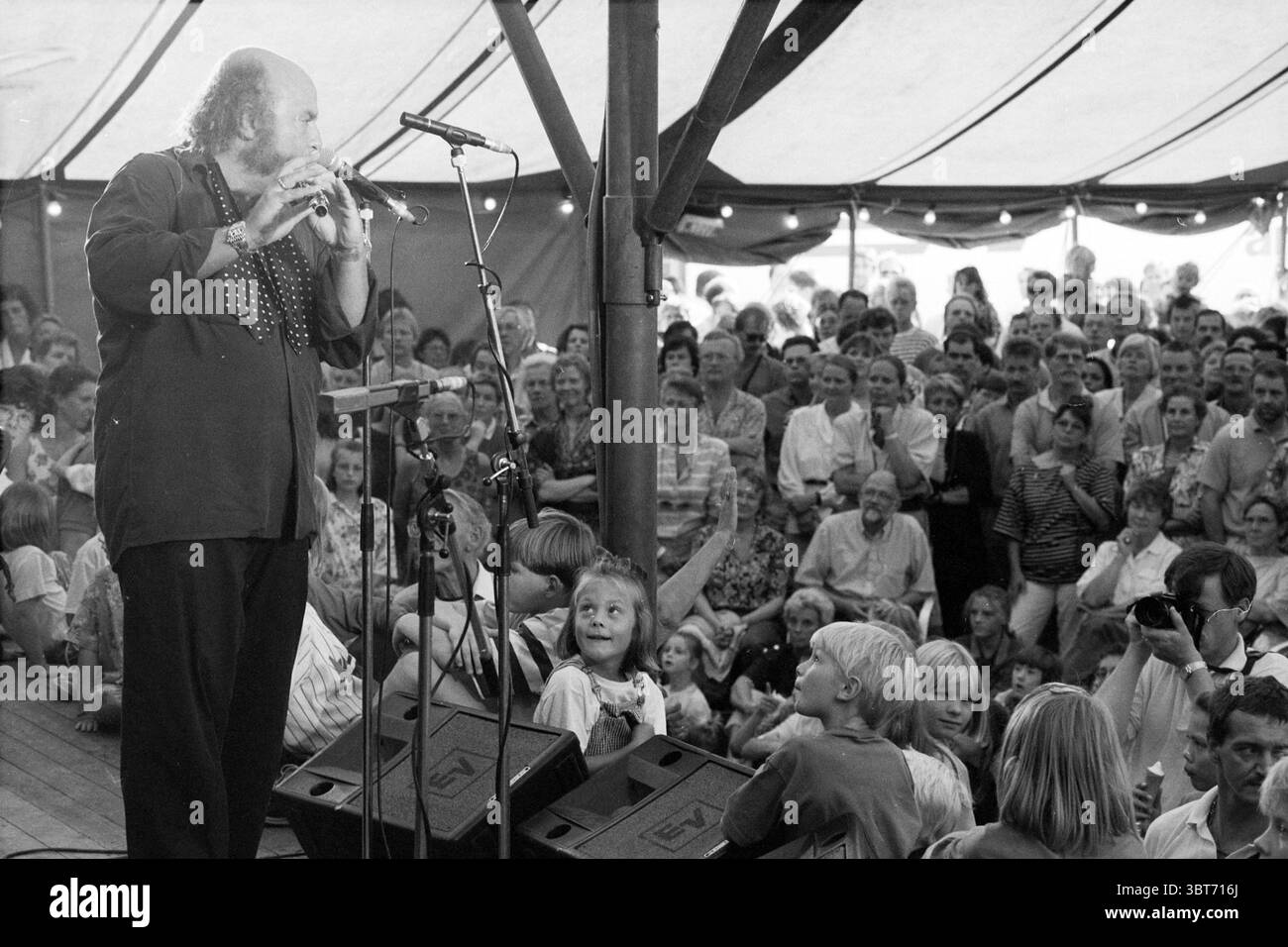 Letzte Aufführung Haarlem Jazz Haarlem Nederland, Whizgle News, Dutch Desk, Niederlande, 1950 - 2000 am 24-08-1991. Das Bild enthält diese Themen. Die Szene spielt in einem großen, lebendigen Zelt voller begeisterter Menschenmengen. Die Atmosphäre ist lebendig und energetisch, spiegelt ein Gefühl der Freude und Aufregung wider. Der Hauptdarsteller, ein Mann mit einem ausgeprägten langen Bart und verspieltem Auftreten, steht an der Spitze, vertieft in das Flötenspiel, was dem Moment eine skurrile Atmosphäre verleiht. Seine Kleidung ist lässig und doch auffällig, geprägt von einem locker sitzenden schwarzen Hemd mit einem auffälligen Muster Stockfoto