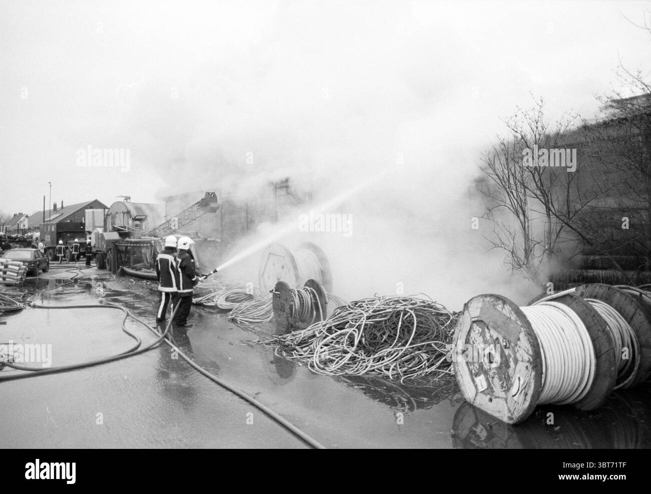 Feuer Tjaden Lager Zuid Schalkwijkerweg Branden Feuerwehr Haarlem Zuid Schalkwijkerweg Niederlande, Whizgle News, Dutch Desk, Niederlande, 1950 - 2000 am 11-03-1989. Das Bild enthält diese Themen. Die Szene zeigt einen Feuerwehrangriff in einem überfüllten Außenbereich, umgeben von dickem weißem Rauch, der nach oben wirbelt und Teile des Hintergrunds verdeckt. Zwei Feuerwehrleute, die mit Schutzausrüstung einschließlich Helmen und Jacken ausgestattet sind, leiten einen starken Wasserstrom aus einem Schlauch aktiv zur Brandquelle. Ihre fokussierten Ausdrücke vermitteln ein Gefühl der Dringlichkeit und Entschlossenheit Stockfoto