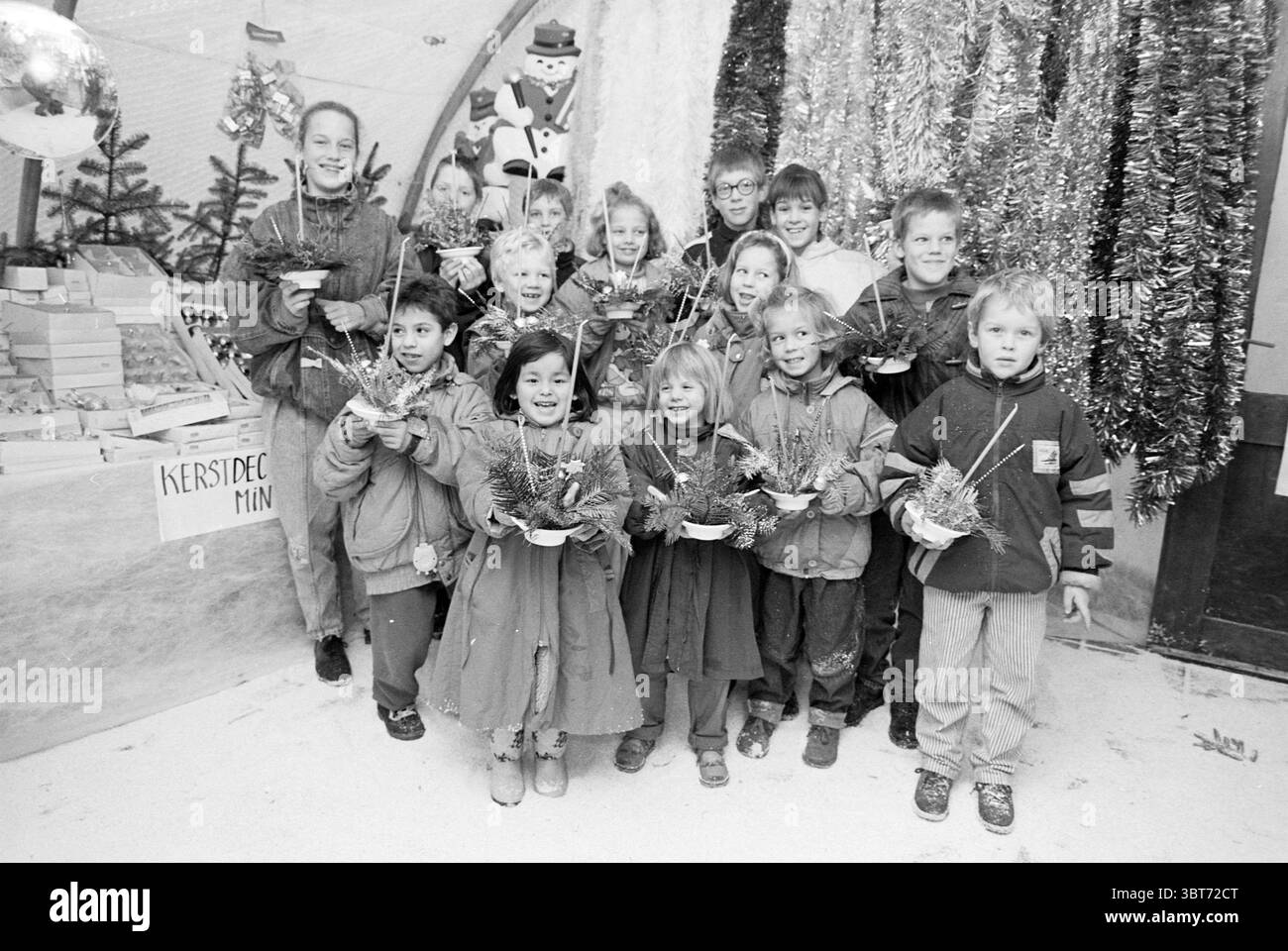 Kinder im Duin Kindergarten wegen Weihnachtsstücken Weihnachten und Weihnachtsbäume Heemskerk, Whizgle News, Dutch Desk, Niederlande, 1950 - 2000 am 13. 12. 1989. Die Abbildung zeigt diese Themen. In einem lebhaften Innenbereich steht eine Gruppe von Kindern vor einer festlichen Kulisse mit Weihnachtsdekoration. Die Szene ist voller Jubel, geprägt von hellen Farben und einer verspielten Atmosphäre. In der Mitte steht ein Mann, wahrscheinlich ein Lehrer oder Hausmeister, mit Stolz, umgeben von einer vielfältigen Gruppe von Kindern, von denen jedes kleine Topfarrangements mit Grün und farbenfrohen Accessoires hält. T Stockfoto